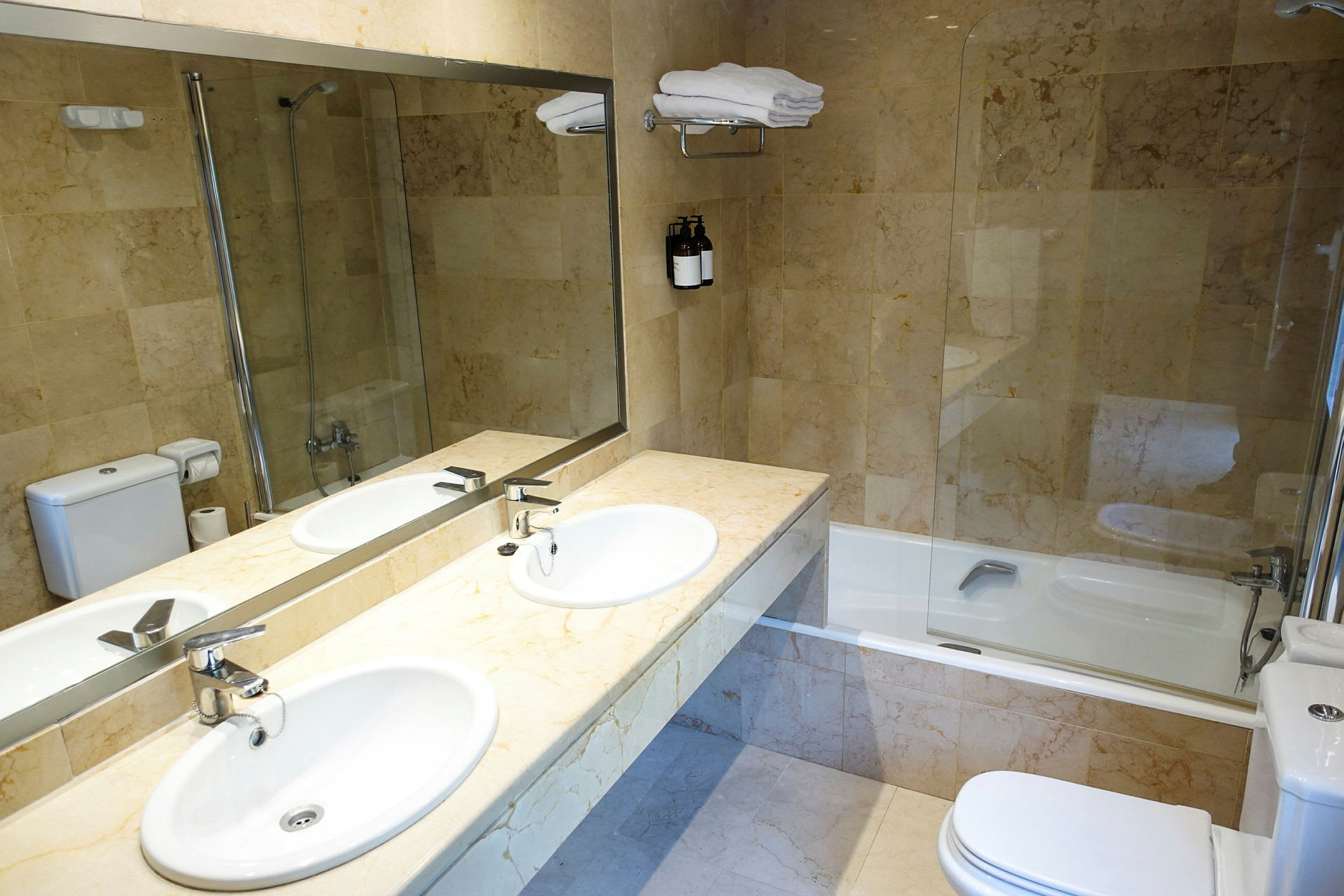Et badeværelse med toilet, badekar med glasvæg, dobbelt håndvask og vægspejl. (AI-genereret tekst)