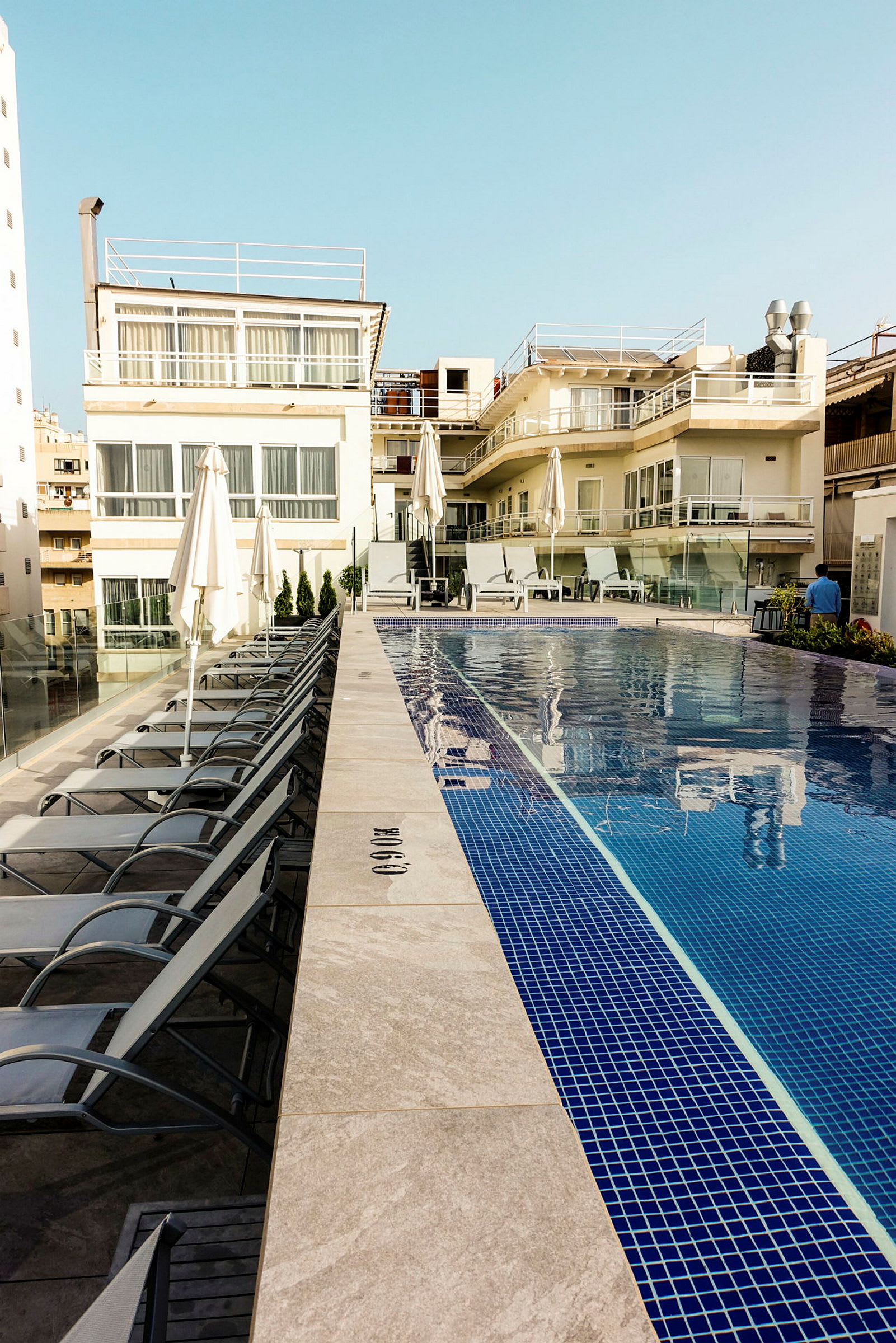 Et poolområde ved et hotel, med liggestole og parasoller opstillet langs poolen foran en bygning. (AI-genereret tekst)
