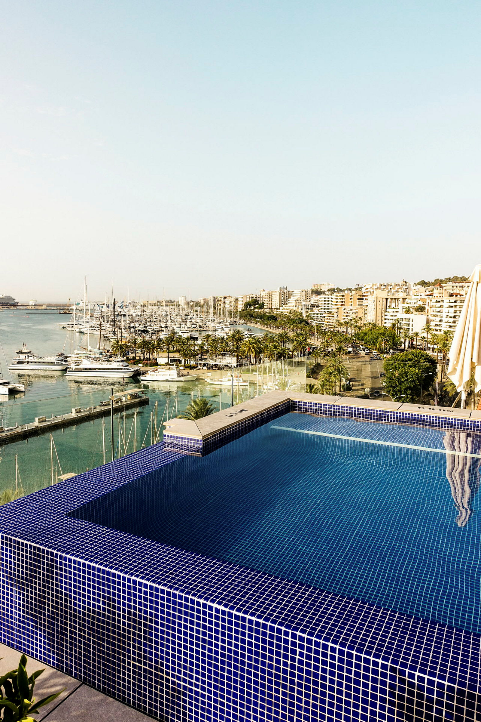 En udsigt over en infinitypool med blå mosaik og en marina med både samt en by i baggrunden. (AI-genereret tekst)