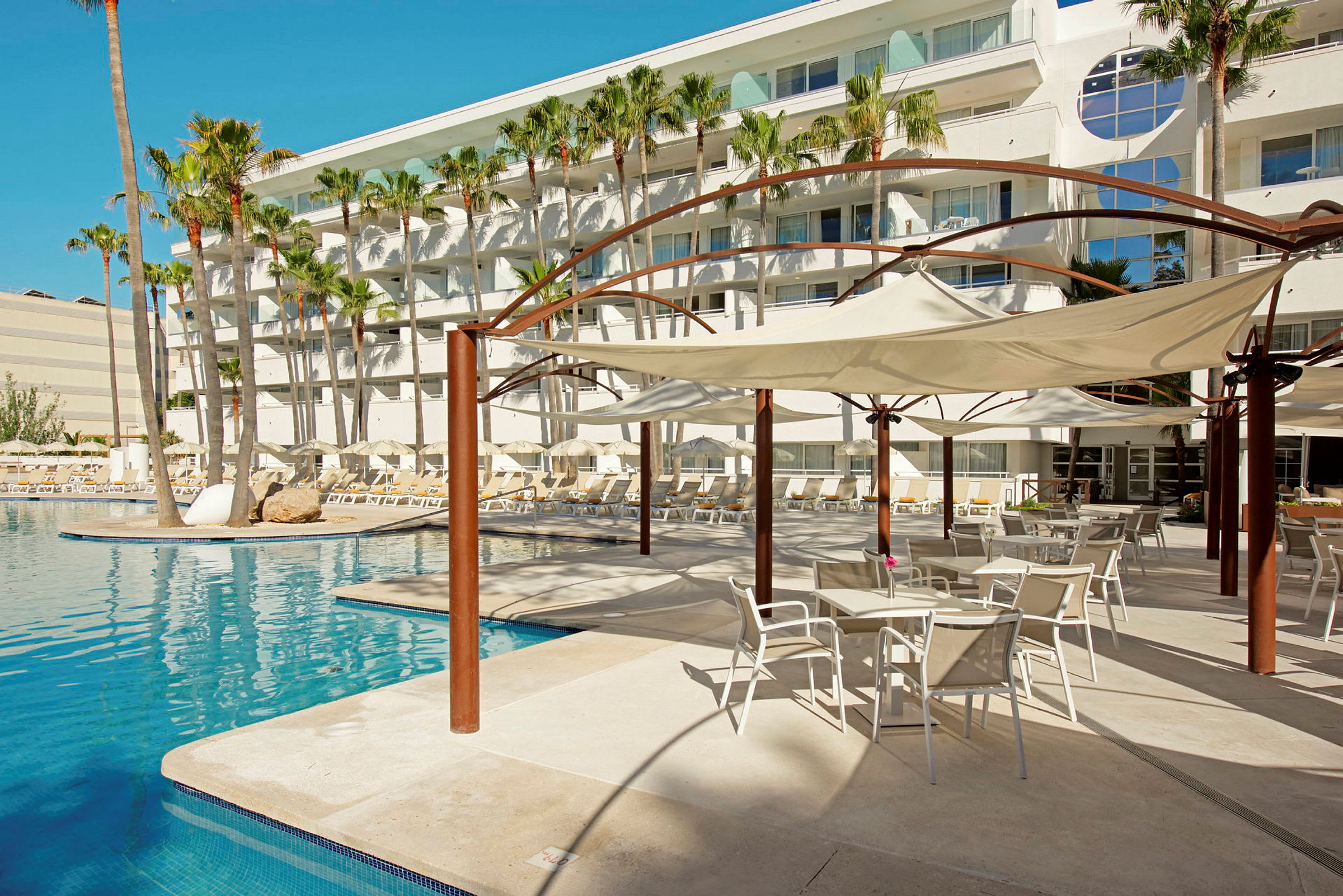 Et hotelområde med en udendørs pool, liggestole og skyggende parasoller. I baggrunden ses et hotel med altaner og omkringliggende palmer. (AI-genereret tekst)
