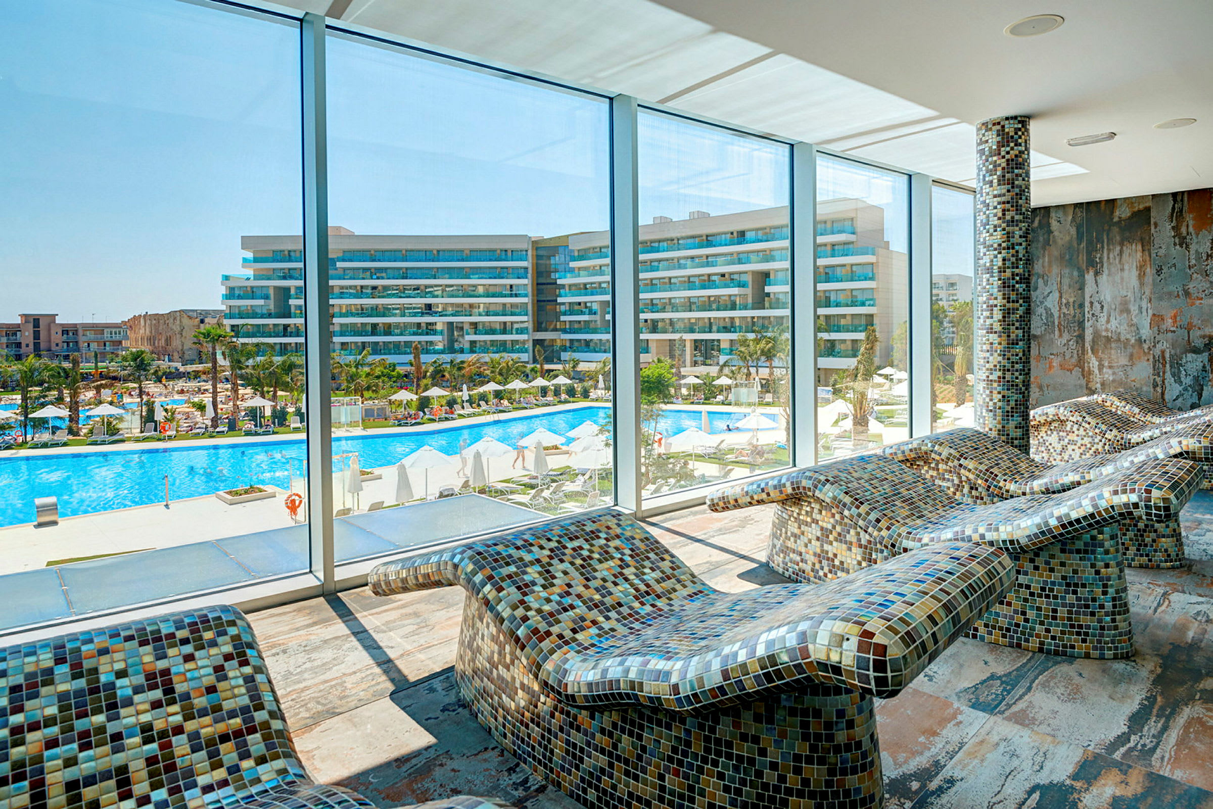 Udsigt fra et rum med mosaikbeklædte liggestole mod en pool og et hotel. (AI-genereret tekst)