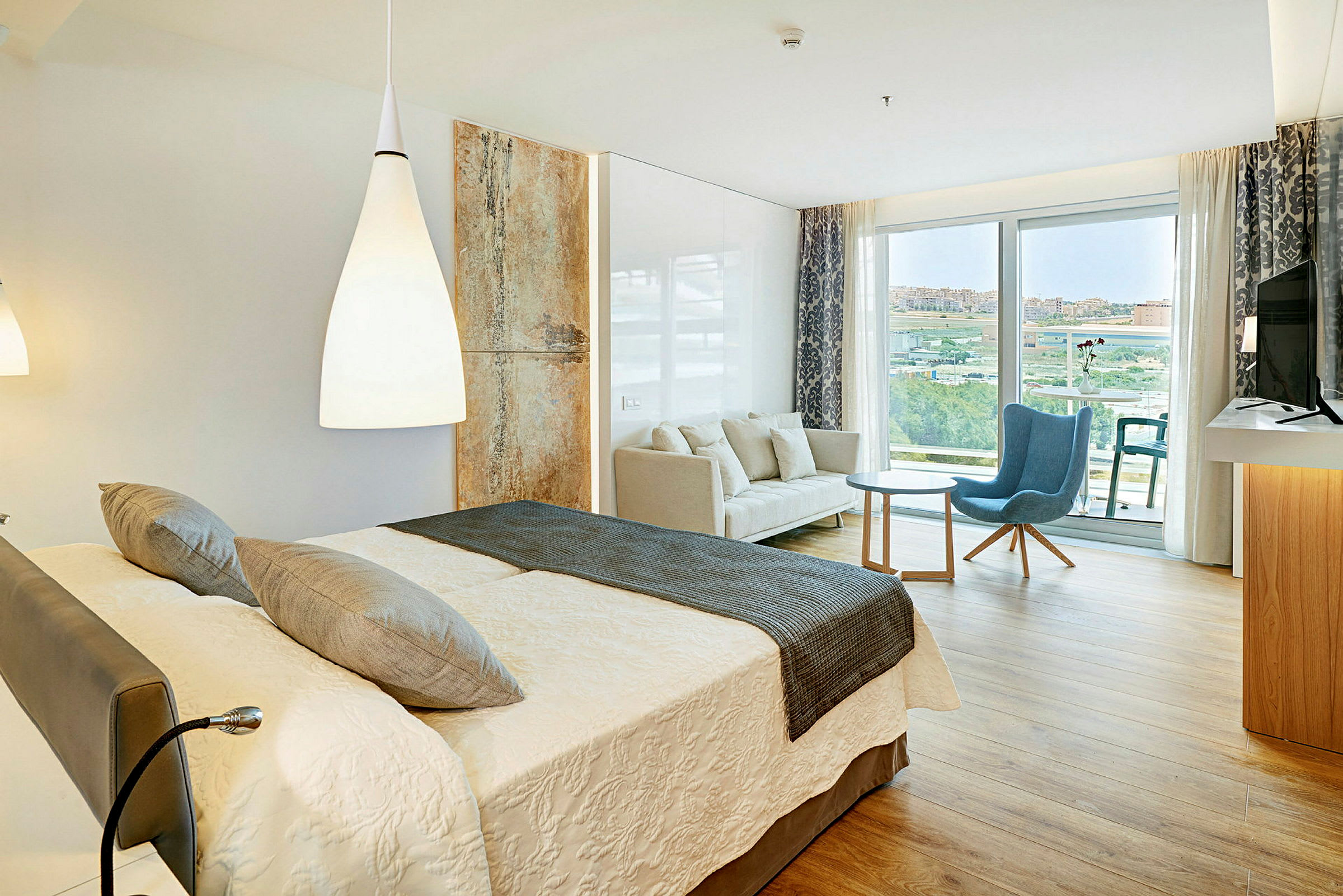 Et hotelværelse med en dobbeltseng, en sofa, en lænestol og et bord samt en balkon med udsigt. (AI-genereret tekst)