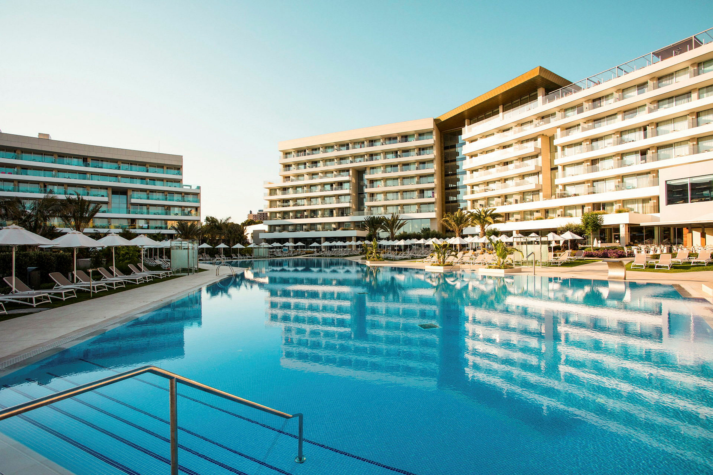 Udendørs pool ved et hotel med liggestole og parasoller, omgivet af bygninger med flere etager. (AI-genereret tekst)