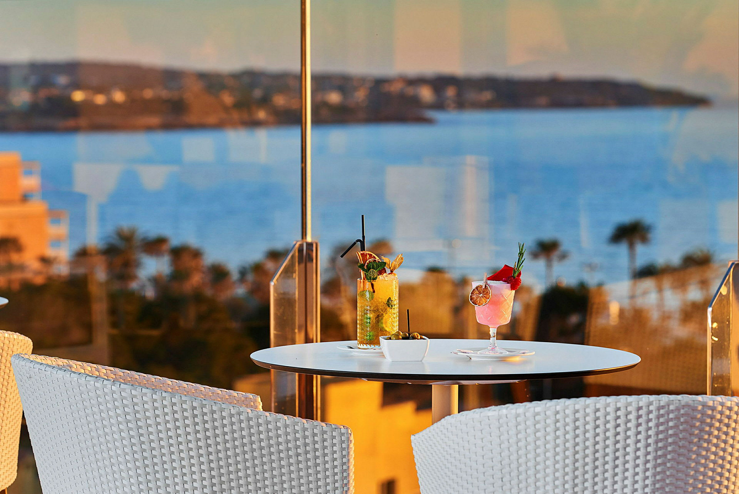 En hvid siddegruppe med et bord, hvorpå der står to cocktails, med udsigt til havet i baggrunden. (AI-genereret tekst)