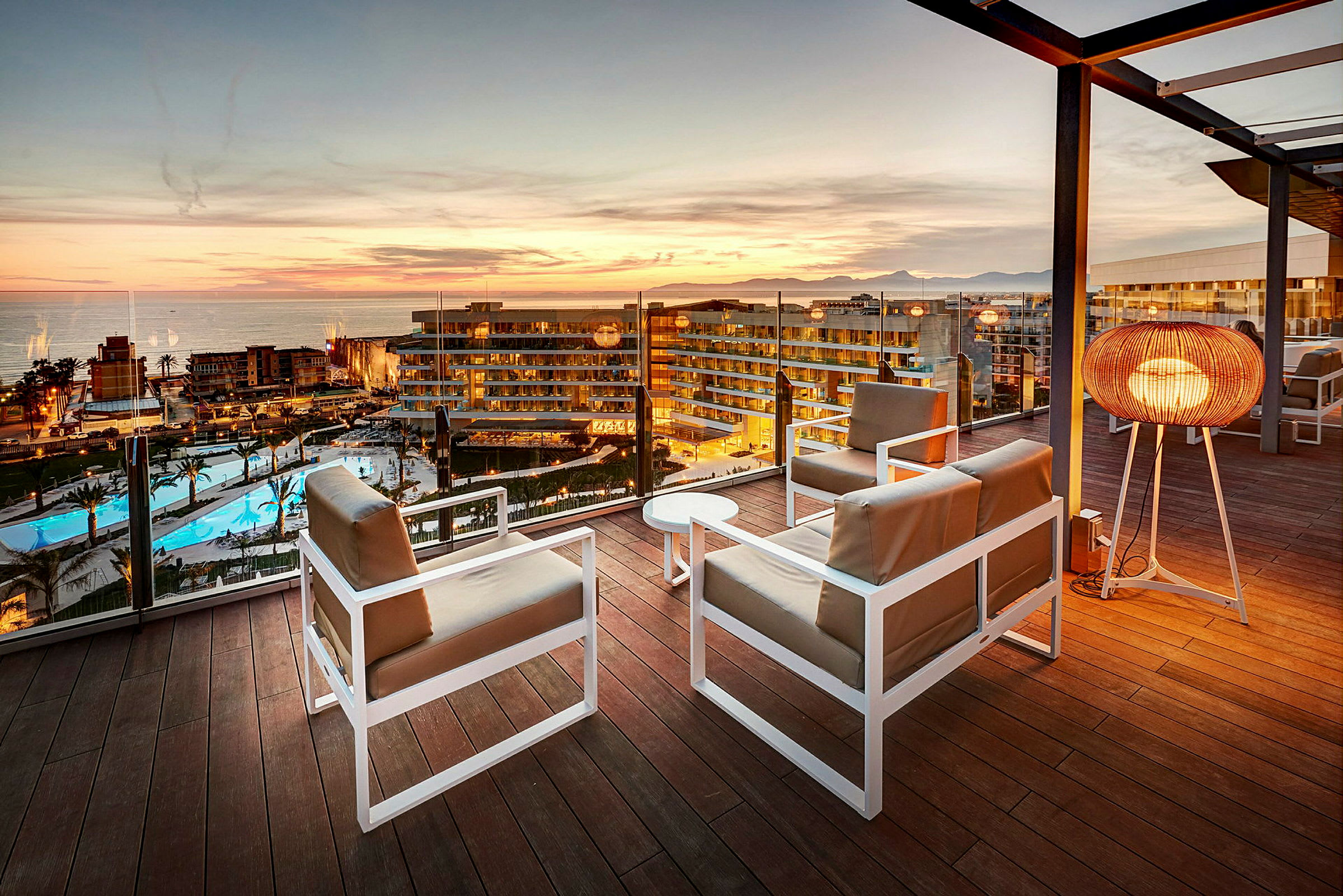 Udsigt over solnedgangen fra en terrasse med siddepladser og en gulvlampe, med hotel og poolområde i baggrunden. (AI-genereret tekst)