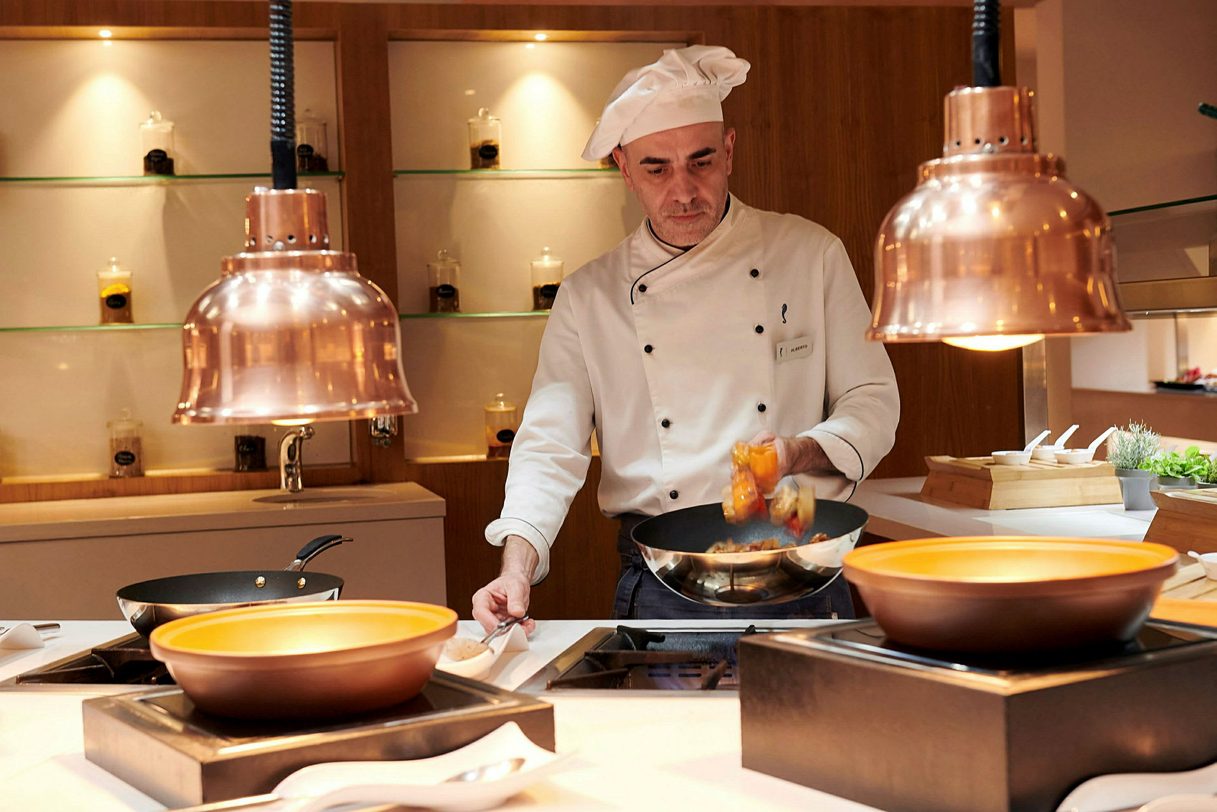 En kok iført hvidt arbejder ved en åben restaurantdisk med kobberlamper ovenover. (AI-genereret tekst)