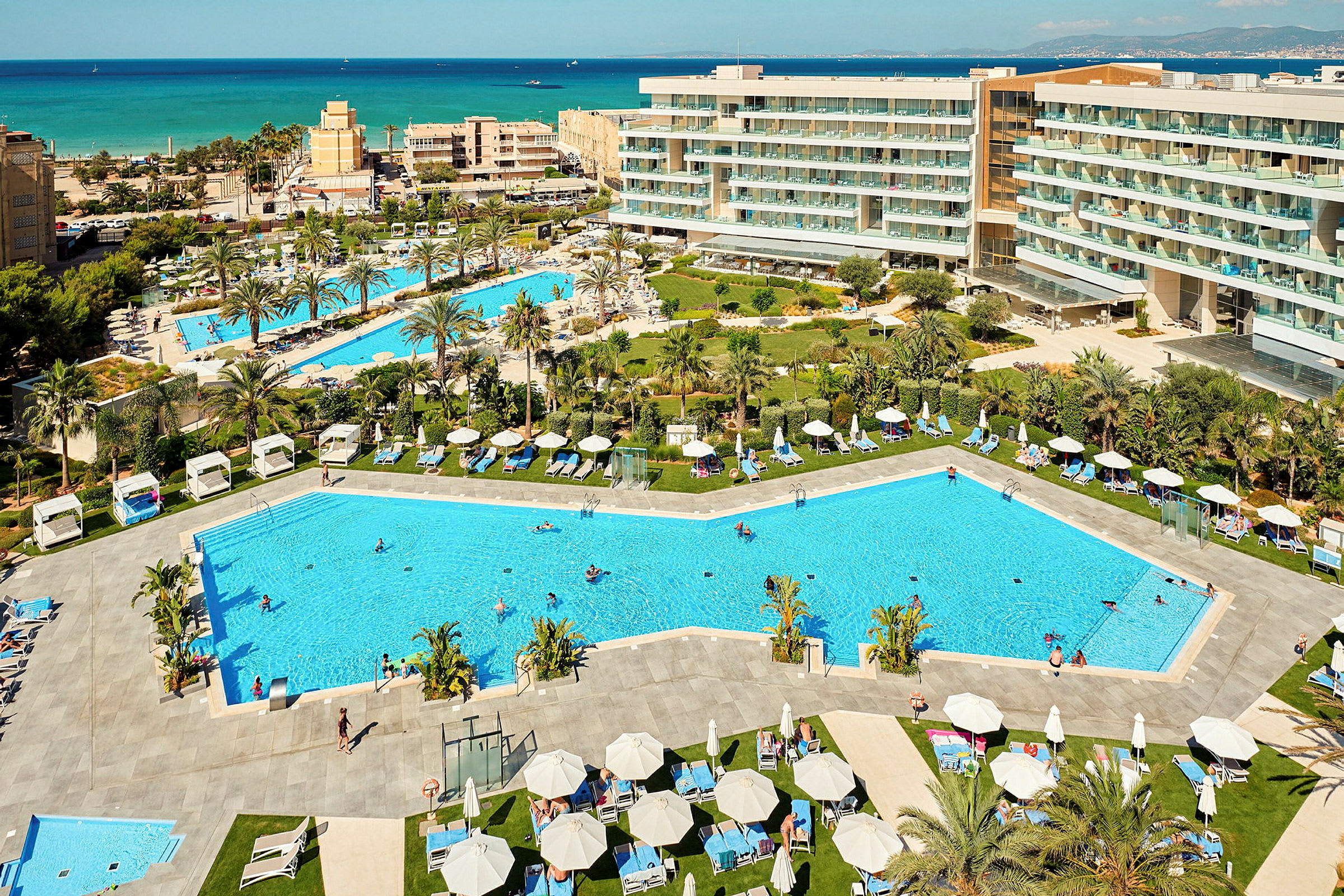 Udsigt over et feriested med swimmingpools, liggestole og omkringliggende hotelbygninger samt havet i baggrunden. (AI-genereret tekst)