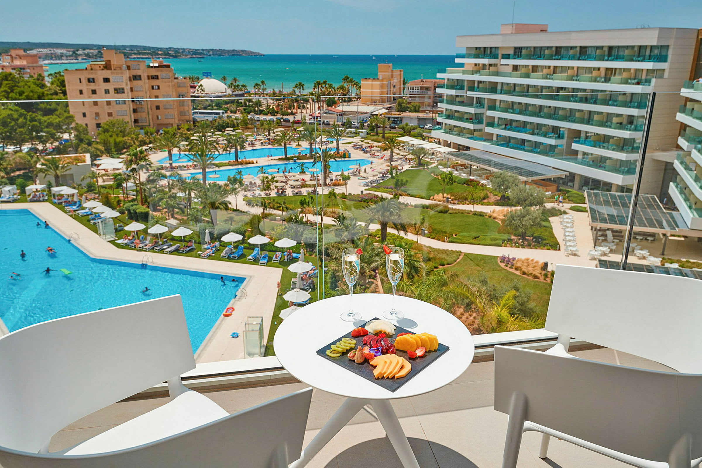 Udsigt fra en balkon mod en hotelpool og havet, med en frugttallerken og to glas på et bord. (AI-genereret tekst)