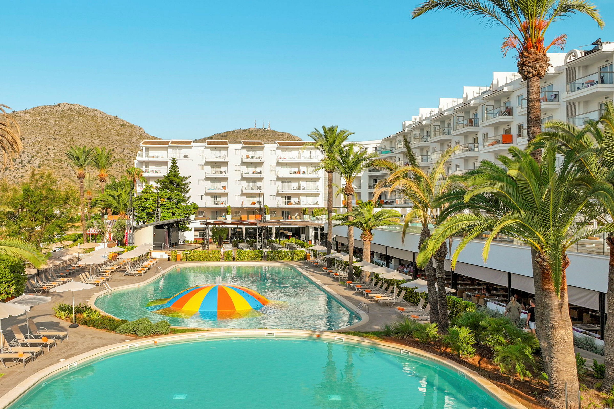 Et hotel med hvide bygninger, liggestole og en pool med en farverig vandstrålende kuppel i midten. (AI-genereret tekst)