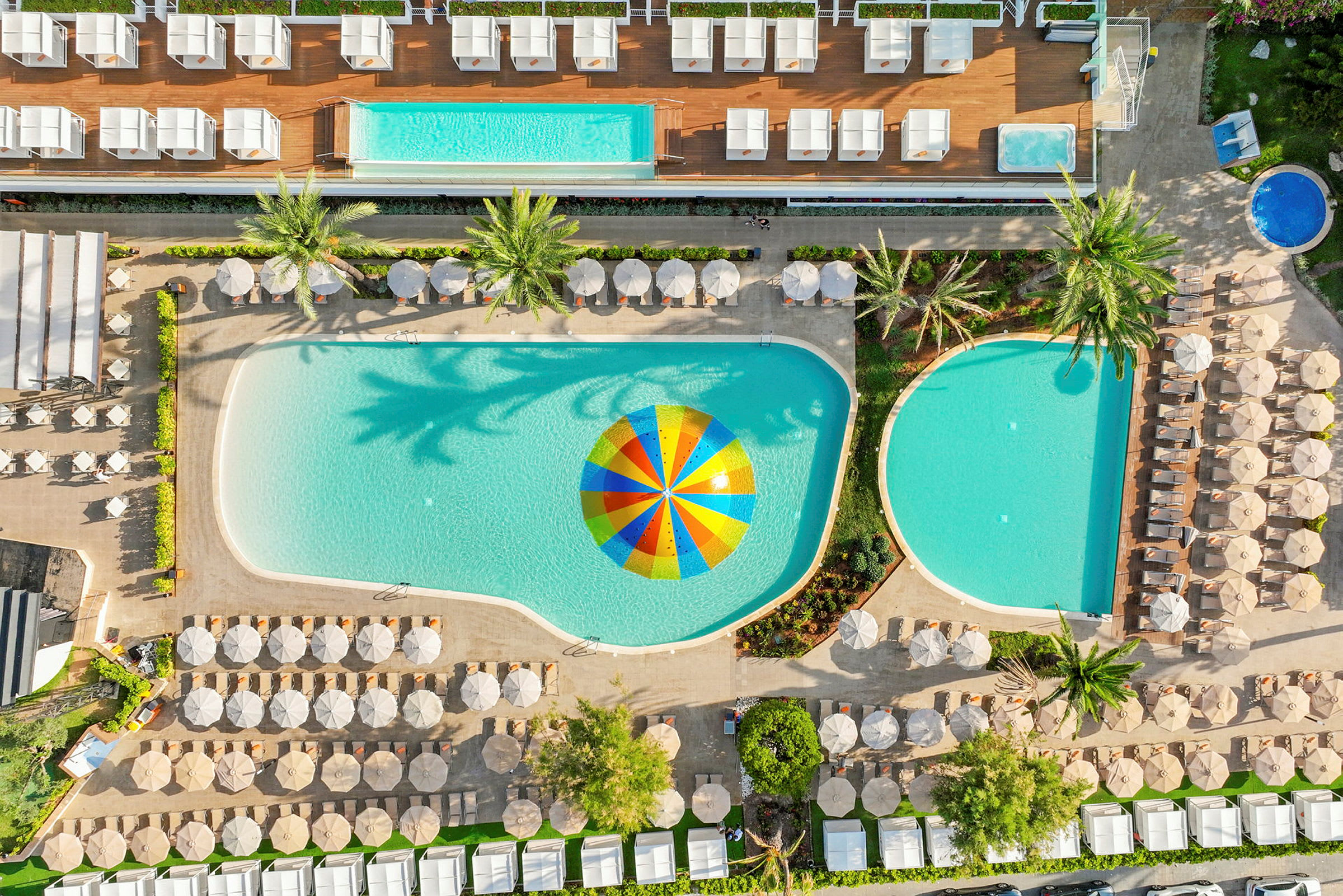 Luftfoto af en hotelpool med parasoller og liggestole omkring samt en farverig parasol i vandet. (AI-genereret tekst)