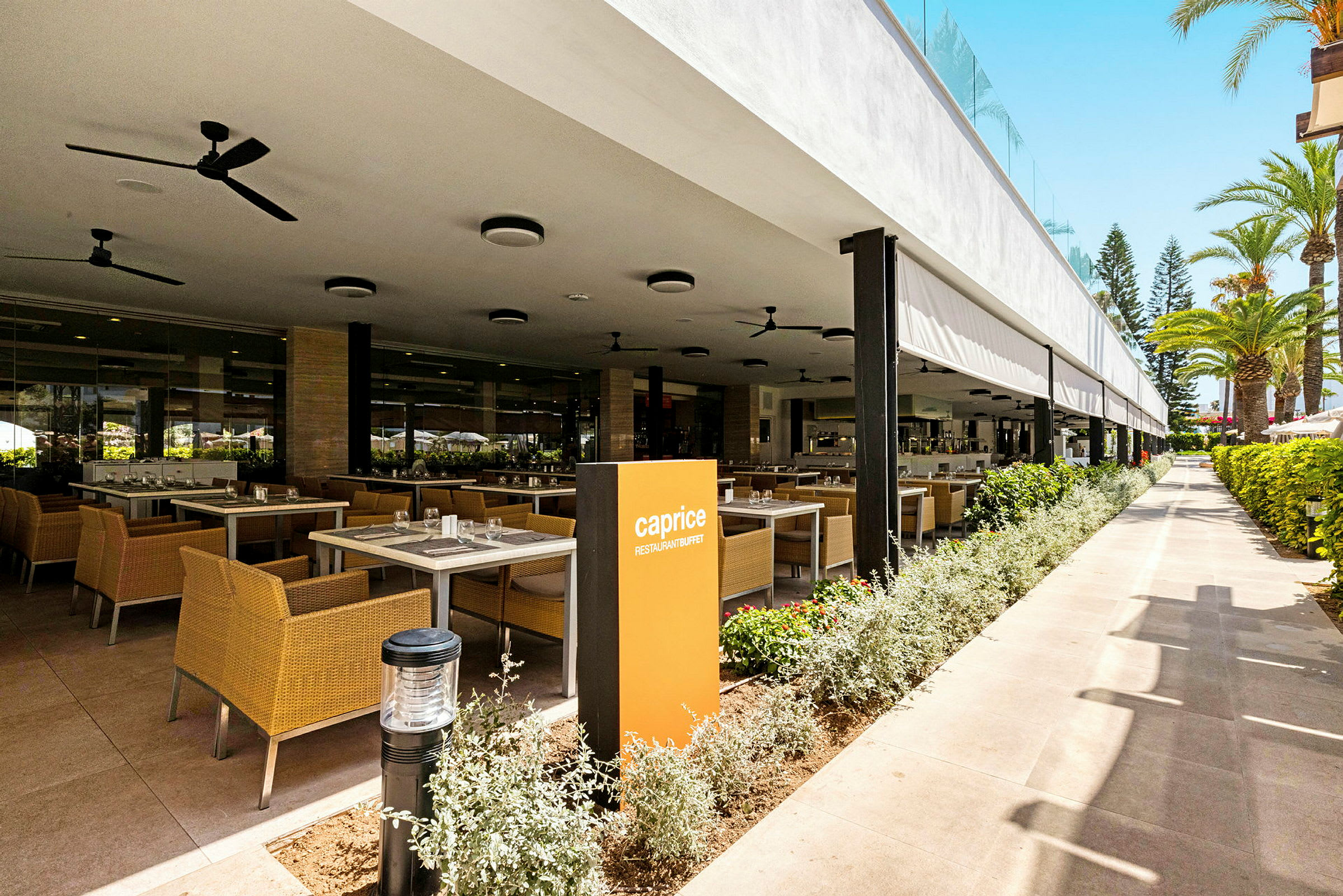 Restaurant med udendørs servering og borde på række nær planter. (AI-genereret tekst)