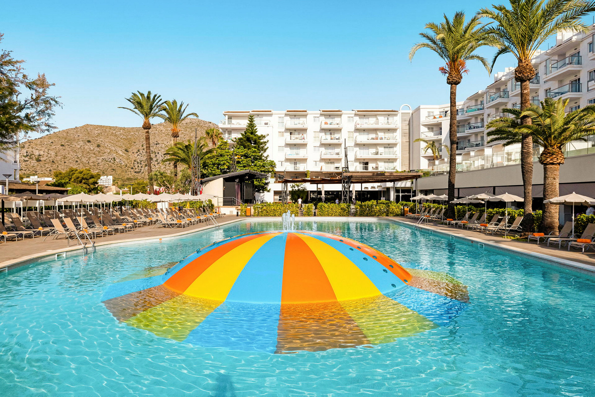 En udendørs swimmingpool med en farverig vandfontæne og omgivende liggestole ved et hotel. (AI-genereret tekst)