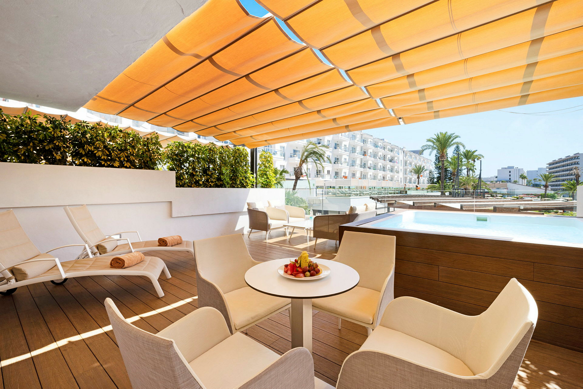 En udendørs terrasse med liggestole, borde og stole. Der er en lille pool og udsigt til hotelbygninger. (AI-genereret tekst)