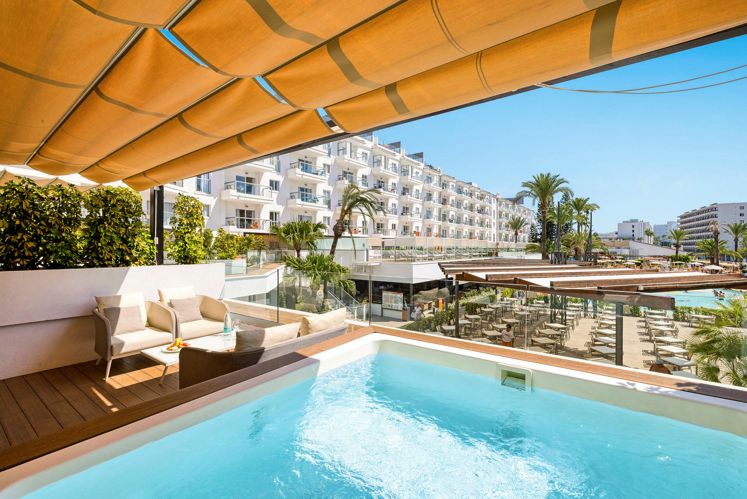 Terrasse med pool, siddegruppe og udsigt over hotelområde med palmer og bygninger. (AI-genereret tekst)