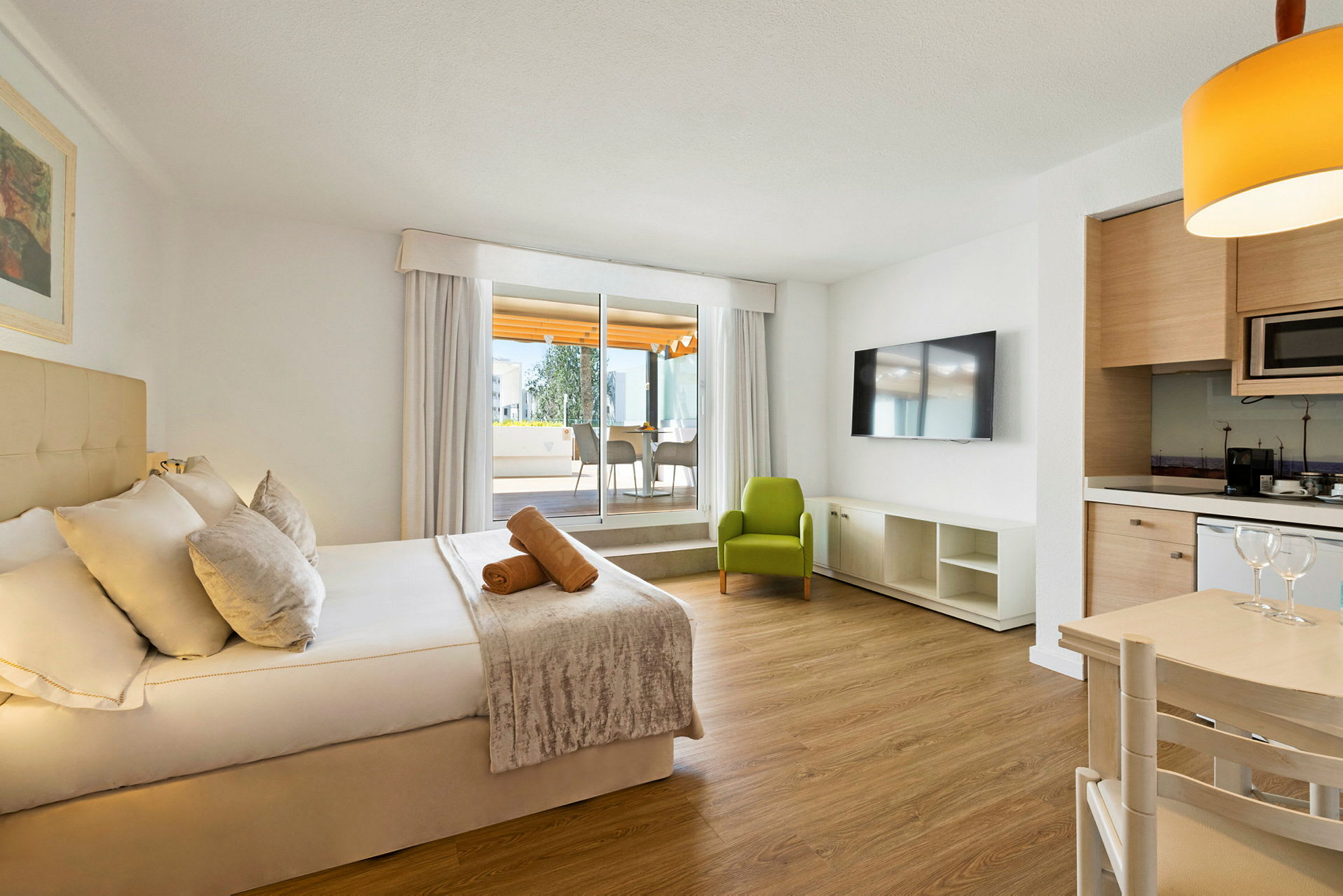 Et moderne hotelværelse med en dobbeltseng, en grøn lænestol, et fjernsyn samt et bord og et køkkenområde. (AI-genereret tekst)