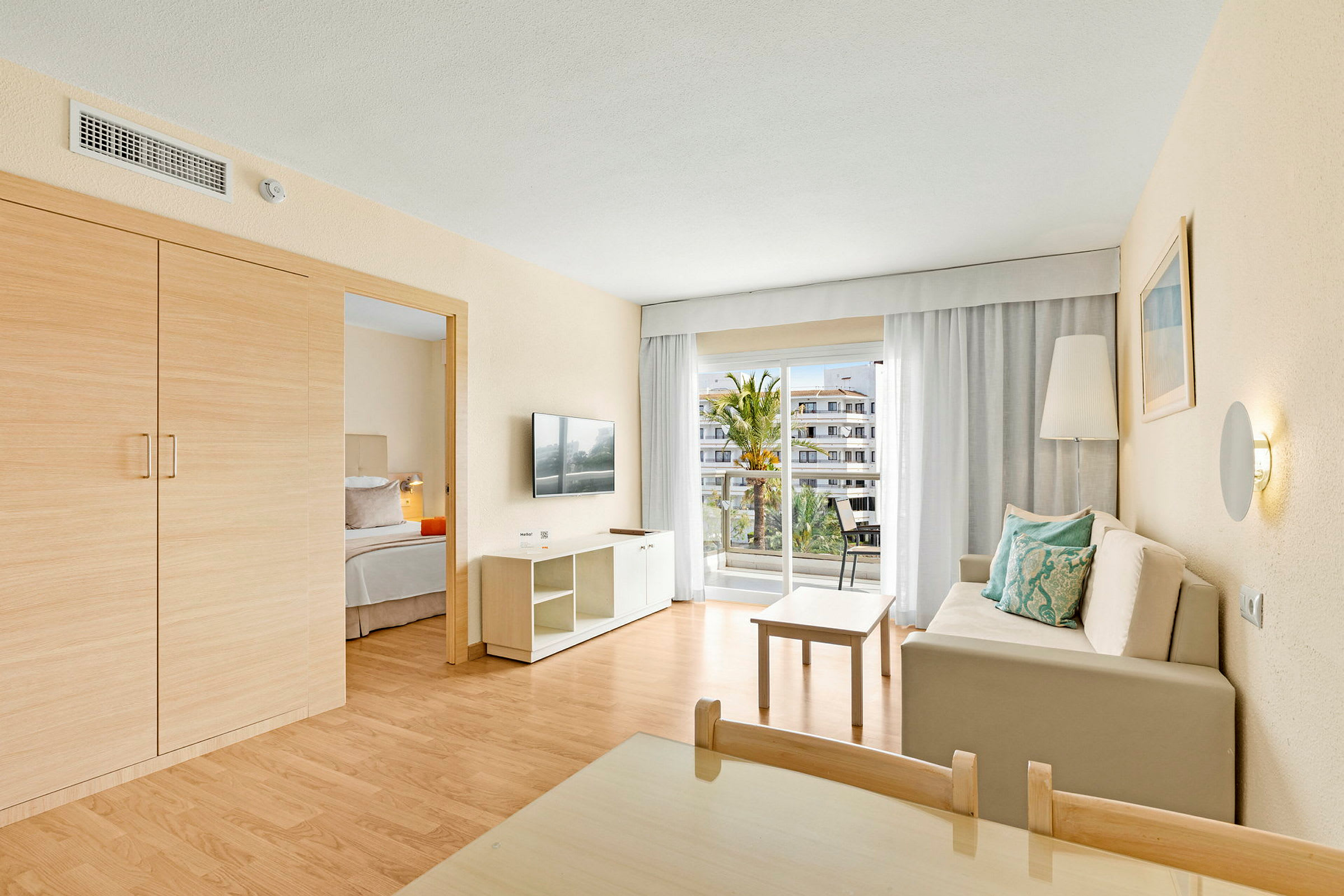 Et hotelværelse med en stue, en sofa med puder, et bord, et TV og en balkon med udsigt. (AI-genereret tekst)