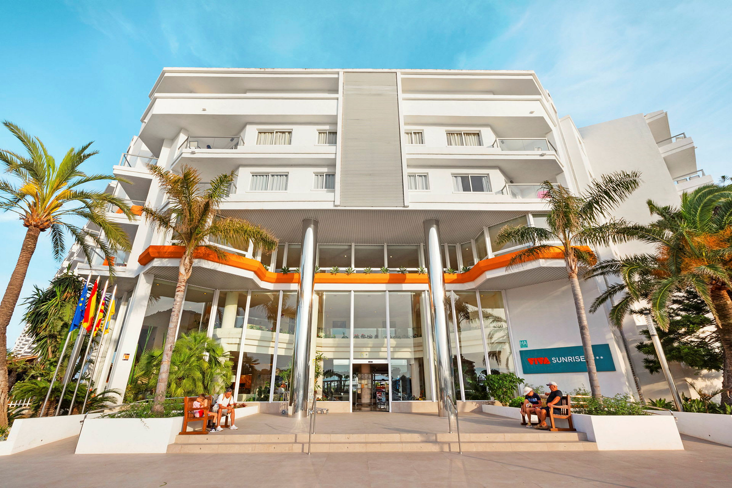 Billede af en moderne hotelbygning med hvid facade, flere etager og store vinduer ved indgangen. Palme og personer ved indgangen. (AI-genereret tekst)