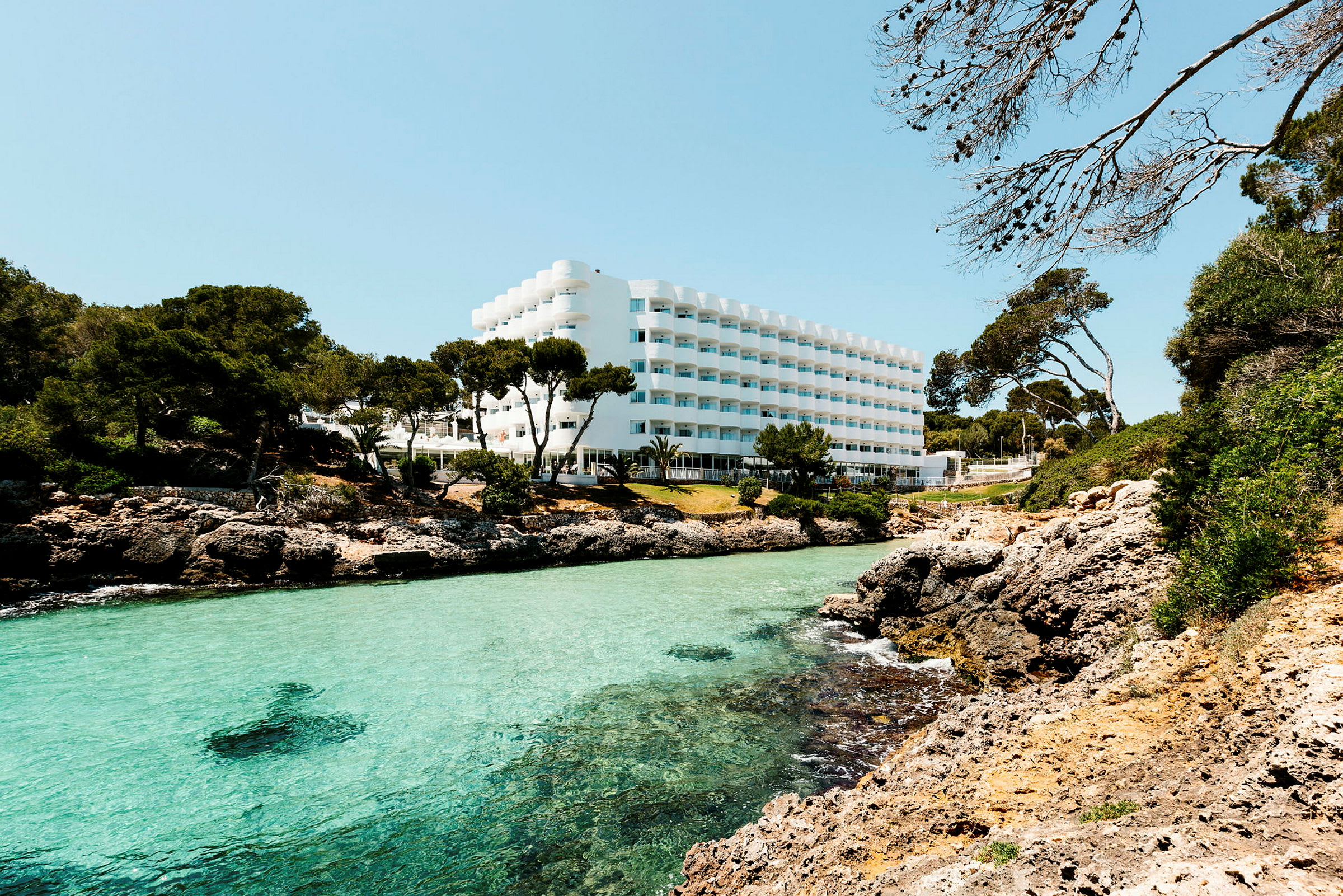 Hotelbygning med hvid facade, beliggende ved en klipperig kyst med klart turkis vand og omgivet af grøntområder. (AI-genereret tekst)