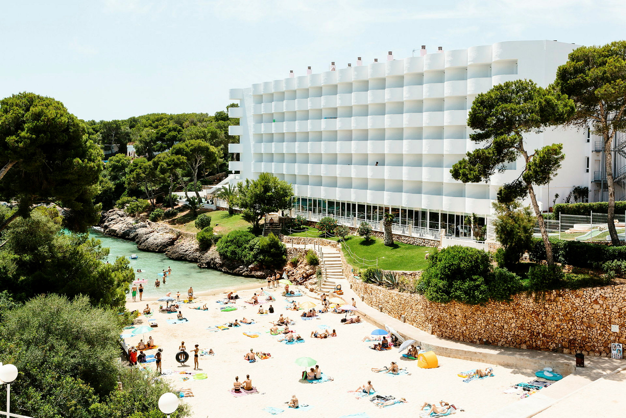 Strand med badende mennesker, omgivet af grønne områder og et hvidt hotel. (AI-genereret tekst)