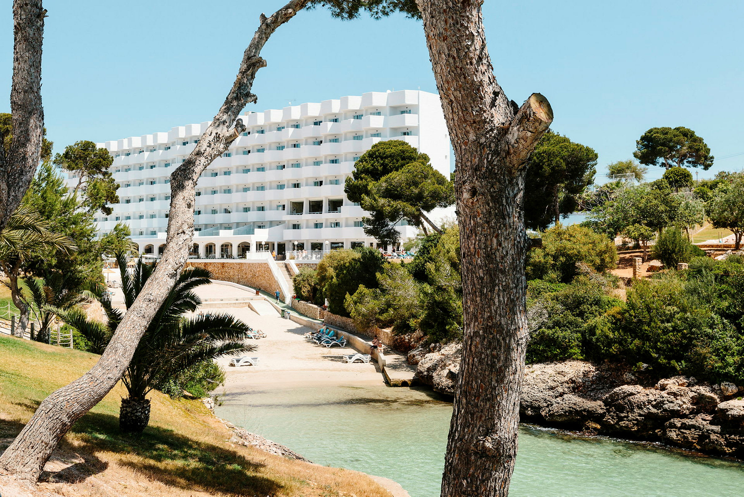 Hotelbygning beliggende ved en lille strand, omgivet af træer og grønne områder. (AI-genereret tekst)