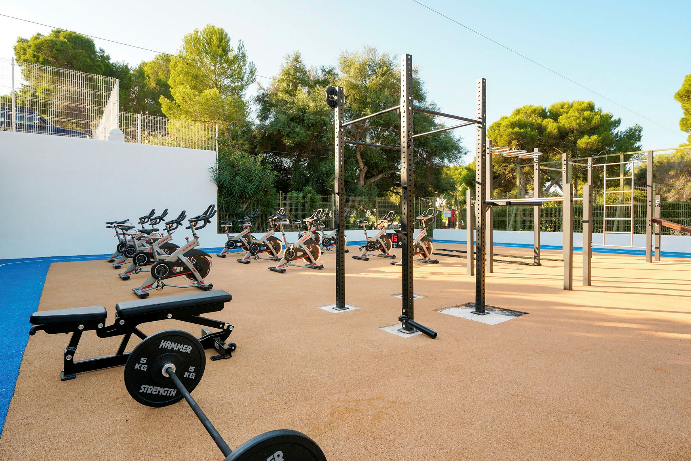 Udendørs fitnessområde med træningsudstyr, herunder motionscykler og vægte. Grøntområder og træer i baggrunden. (AI-genereret tekst)