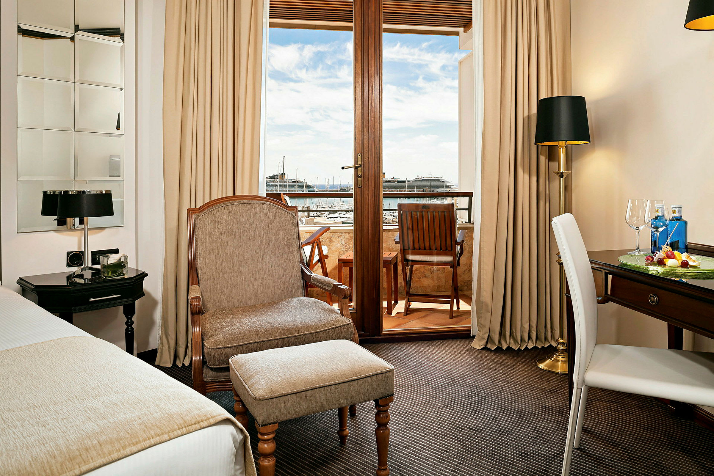 Et hotelværelse med en lænestol, et skrivebord og udsigt til en balkon med siddeplads samt en havn i baggrunden. (AI-genereret tekst)
