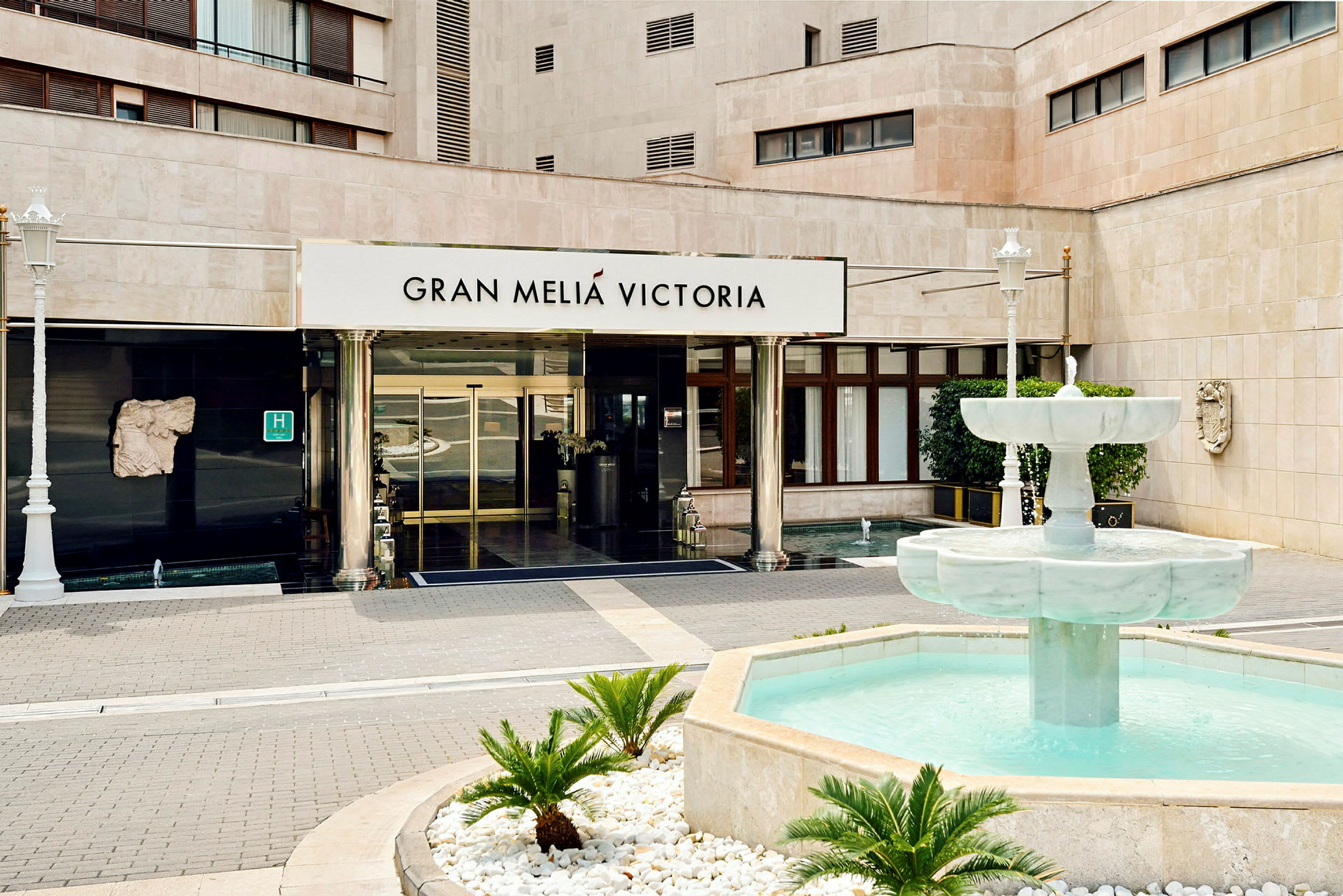 Indgangen til en bygning med teksten "Gran Meliá Victoria" over dørene og en springvand med klart vand foran. (AI-genereret tekst)
