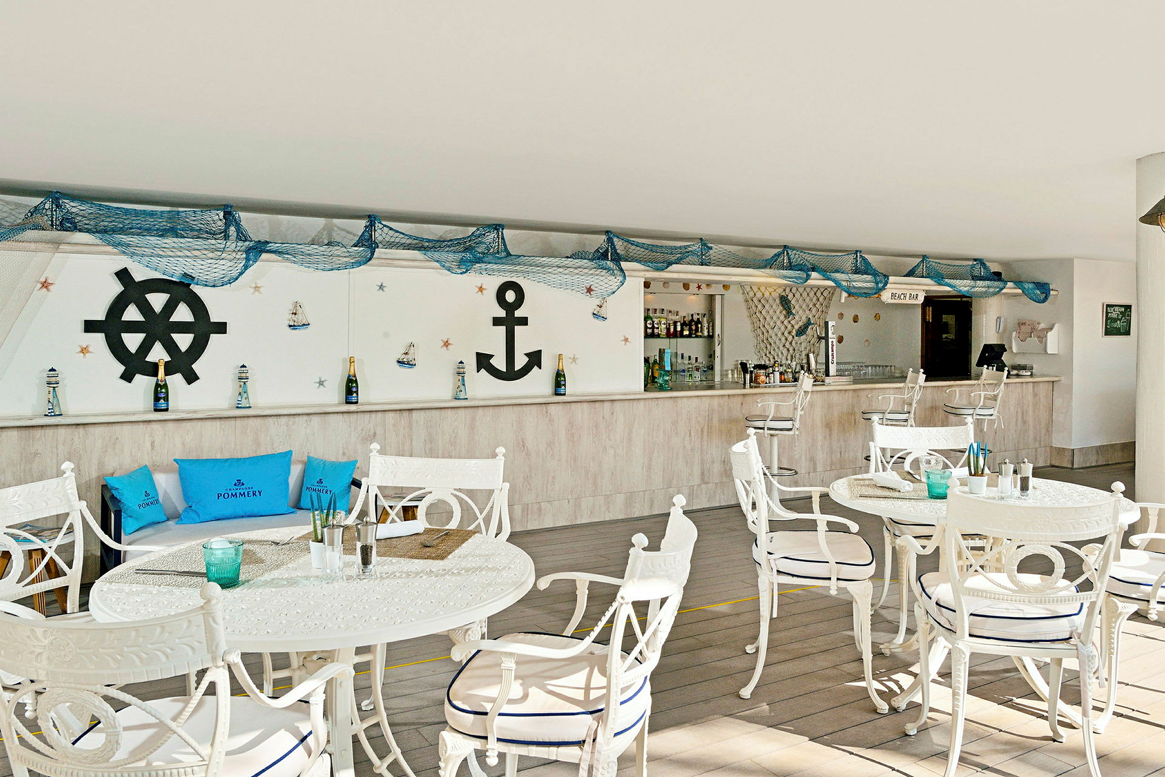 Billede af en strandbar med maritim indretning, herunder hvide borde og stole, blå puder og dekorationer som ankre og fiskernet. (AI-genereret tekst)