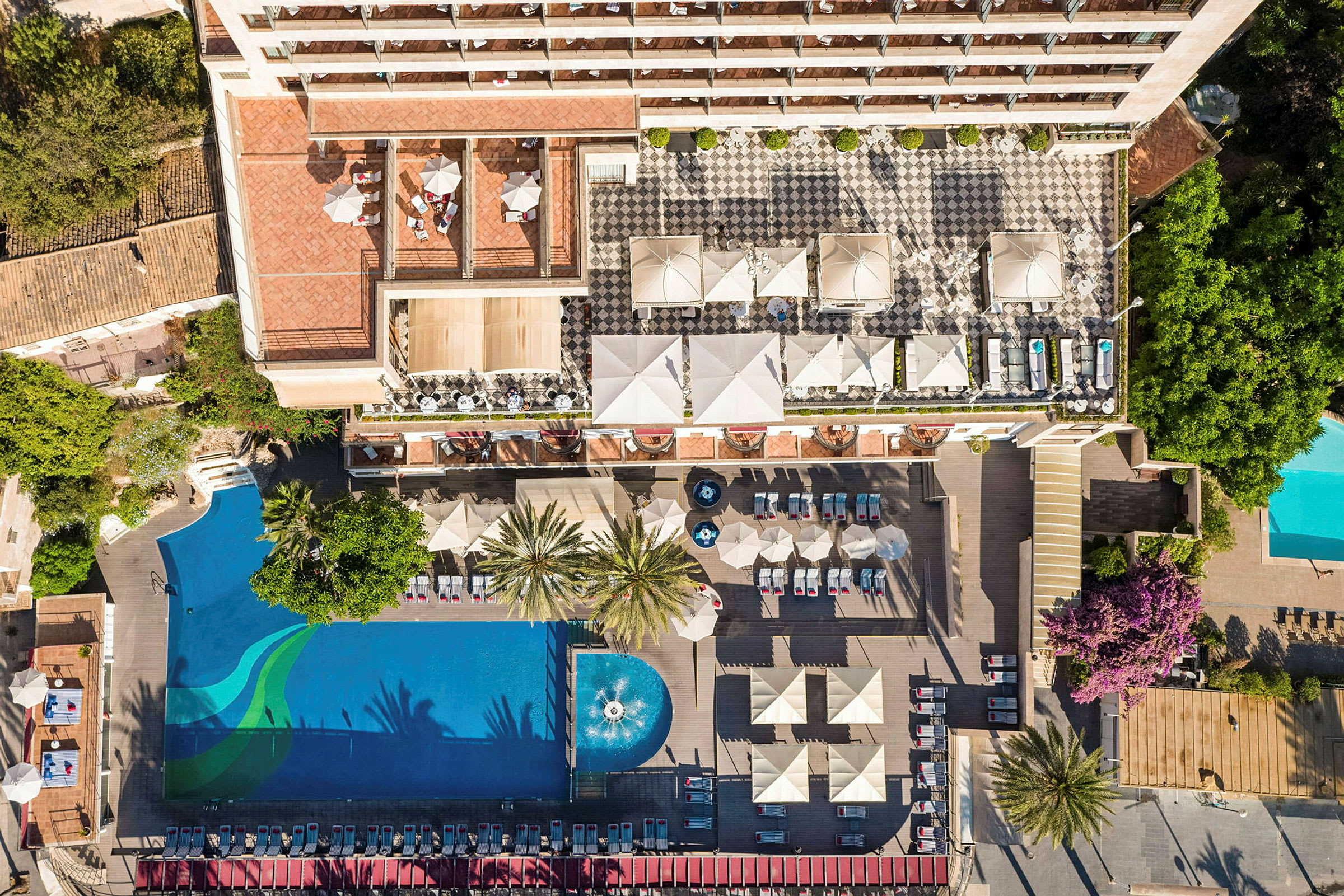 Dronefoto af en hotelkompleks med en pool, liggestole, parasoller og en terrasse. (AI-genereret tekst)