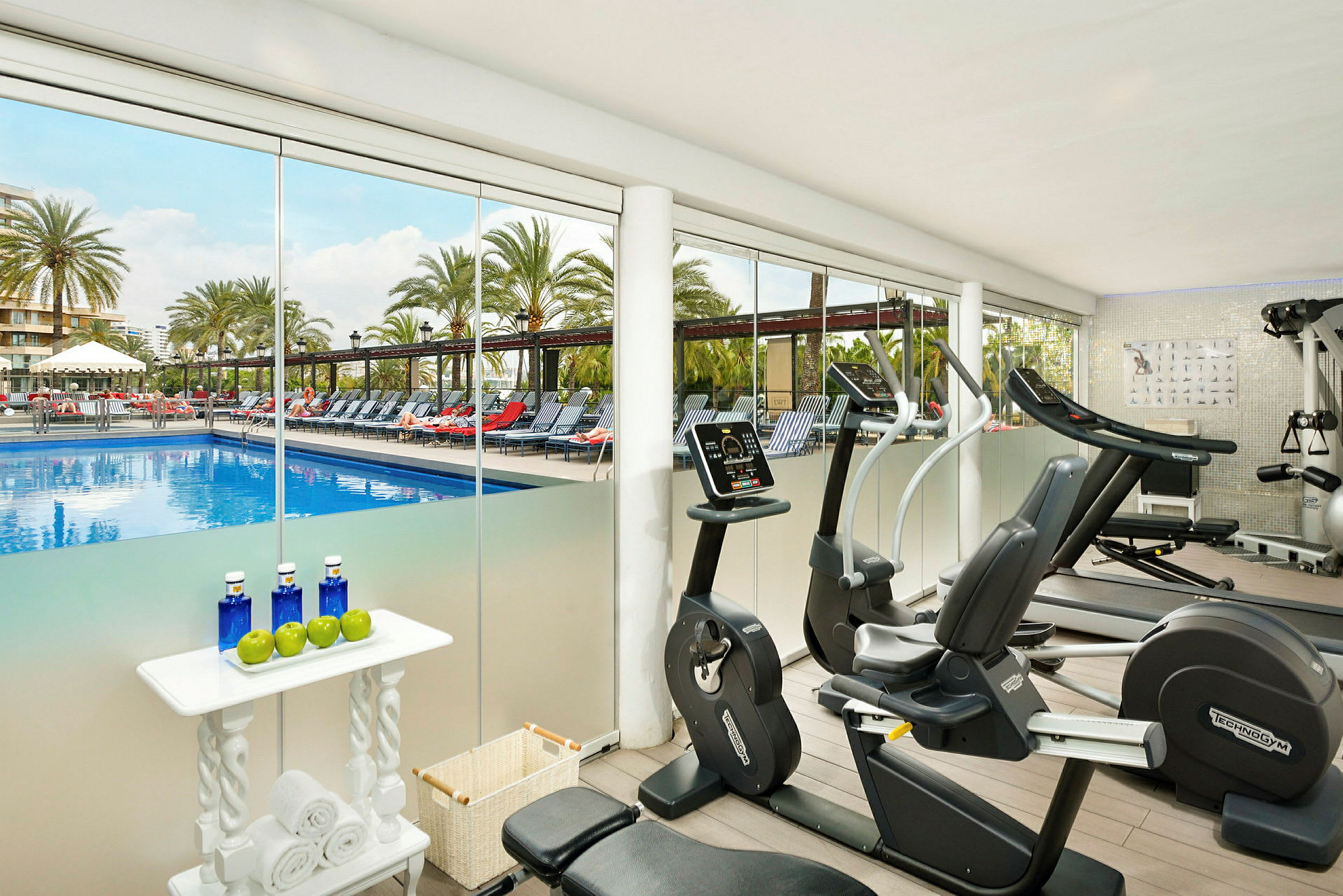 Et fitnesscenter med træningsmaskiner og udsigt over et poolområde med liggestole og palmer. (AI-genereret tekst)