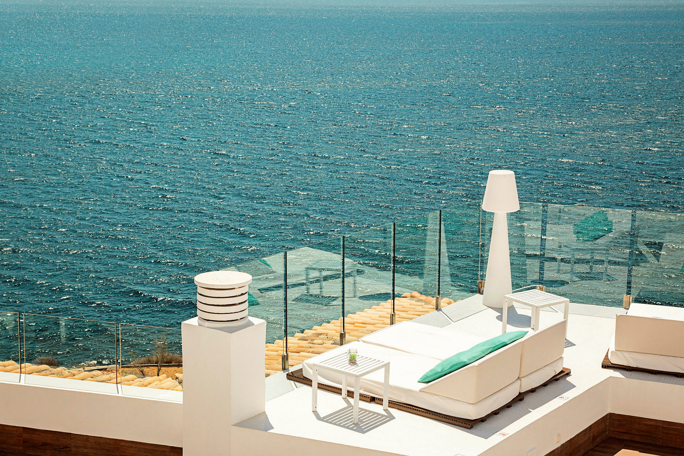 Udsigt over havet fra en terrasse med hvide møbler og glasrækværk. (AI-genereret tekst)