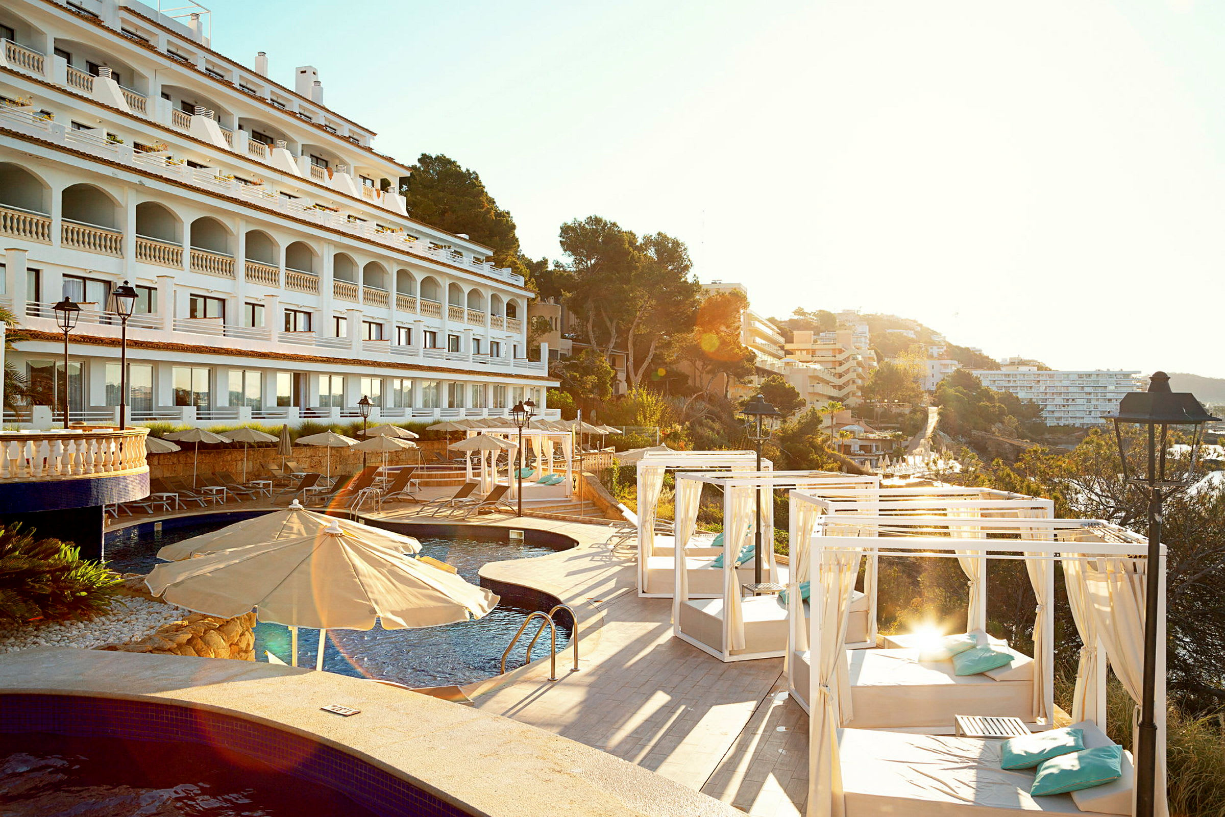 Hotellets facade i hvidt med arkader, swimmingpool og liggestole med parasoller og dagsenge, omgivet af træer og bygninger. (AI-genereret tekst)