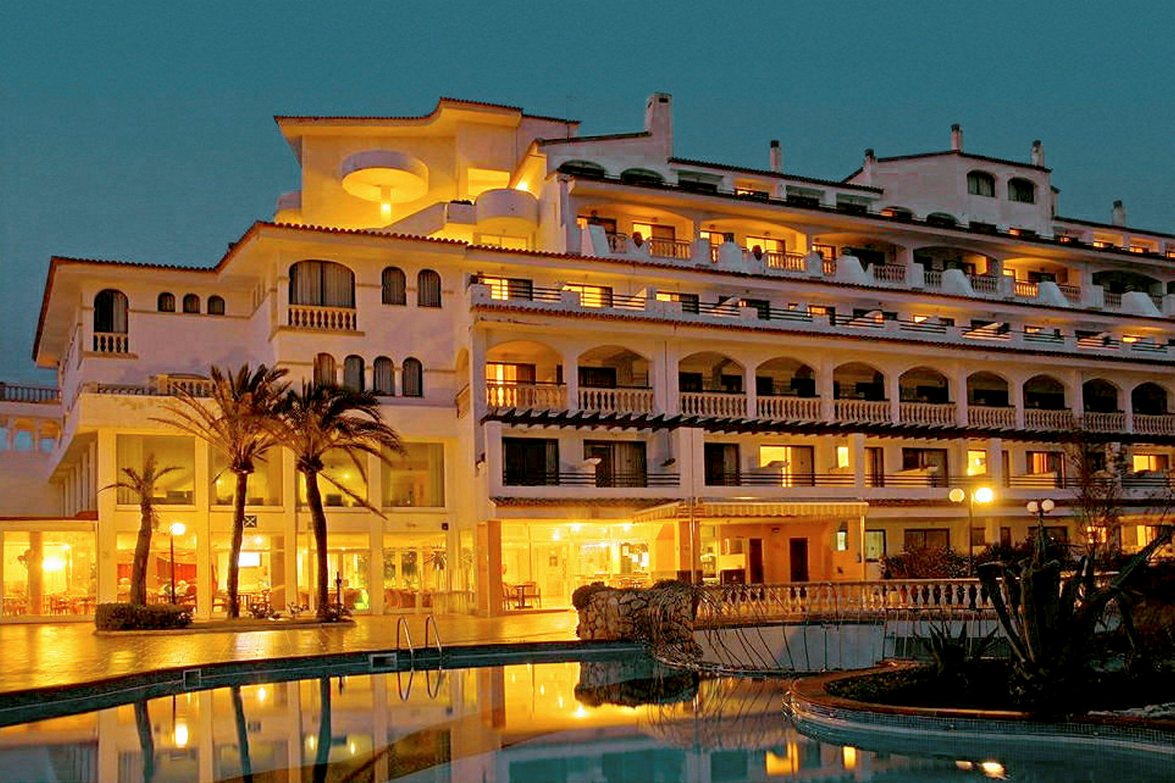 Et fleretasjes hotel med oplyste altaner og palmer foran en pool. (AI-genereret tekst)