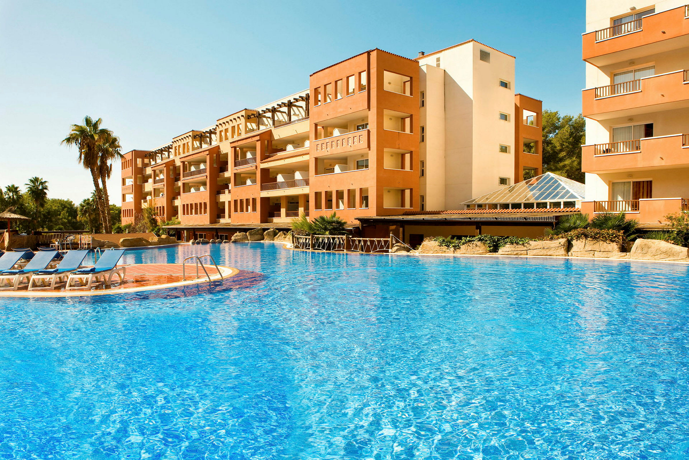 Billedet viser en bygning ved en stor udendørs pool med blåt vand og liggestole placeret ved poolkanten. I baggrunden ses nogle palmer. (AI-genereret tekst)