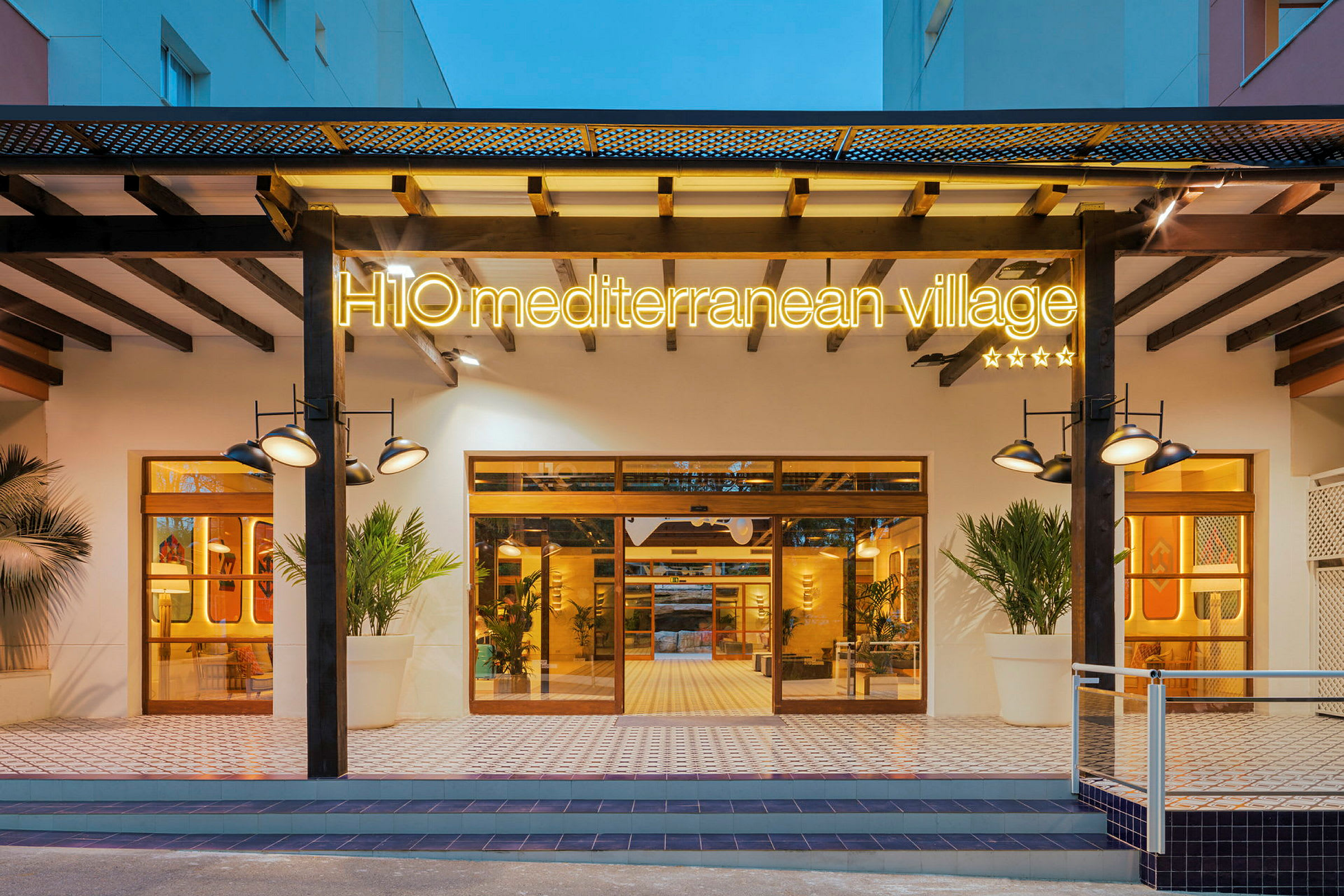 Indgangen til H10 Mediterranean Village, et hotel med oplyst skilt, store vinduer og moderne design. (AI-genereret tekst)