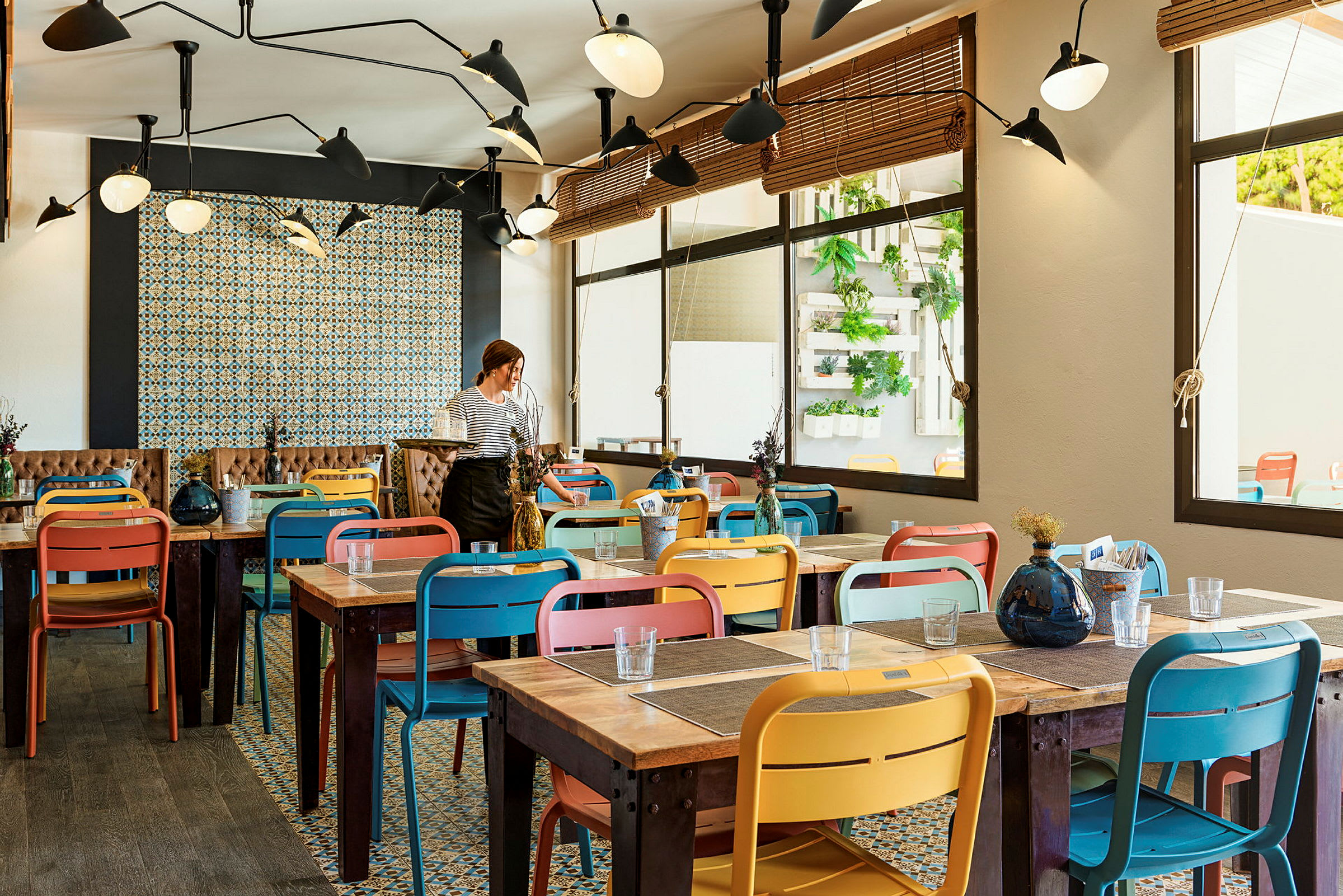 Indendørsbillede fra en restaurant med farverige stole og træborde. En person står ved et af bordene. (AI-genereret tekst)