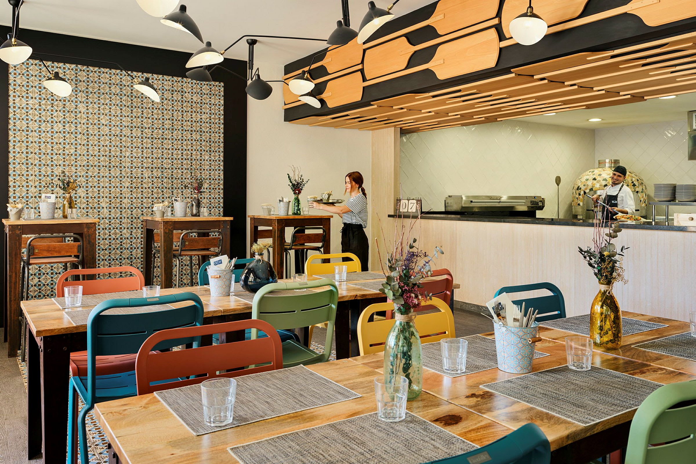 Billede af en restaurantindretning med farverige stole og træborde, dekoreret med blomster i vaser. Væggene har mønstret fliser, og køkkenet er synligt i baggrunden. (AI-genereret tekst)