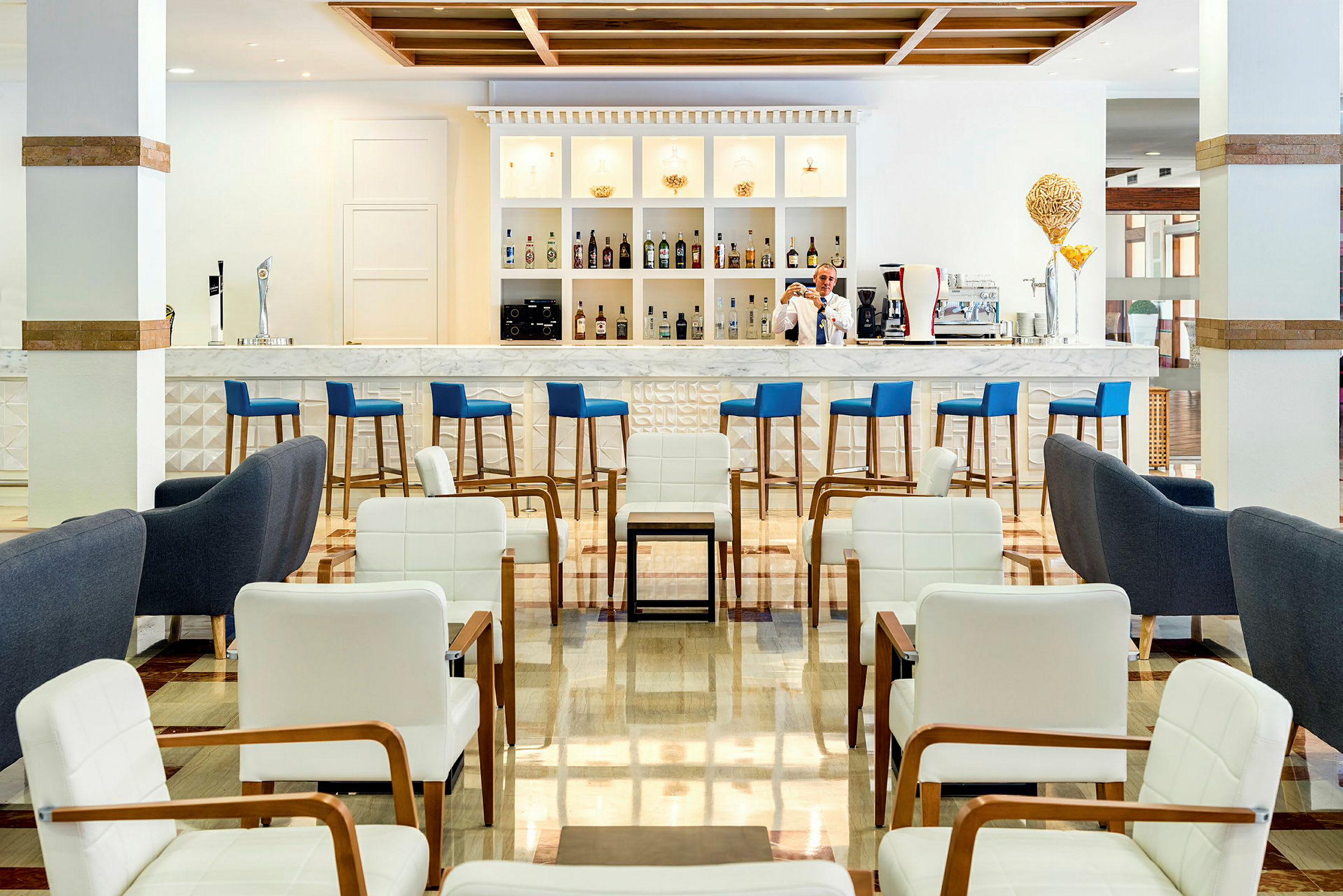 En moderne hotelbar med blå barstole, marmordisk og hvidt interiør. Foran er en siddegruppe med hvide og grå lænestole. (AI-genereret tekst)