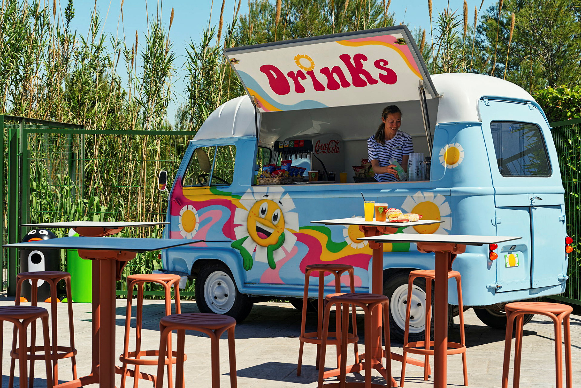 En farverig madbil med teksten 'Drinks' serverer drikkevarer og snacks. Foran bilen står borde og stole. (AI-genereret tekst)