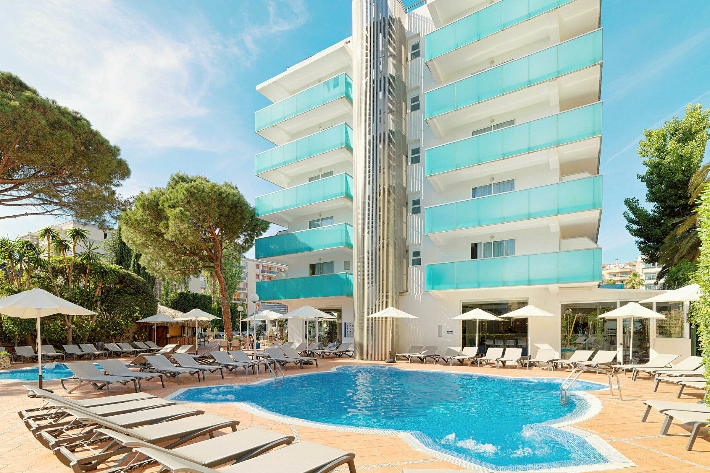 Lejlighedsbygning med balkoner i turkis glas ved en rund swimmingpool omgivet af liggestole og parasoller. (AI-genereret tekst)