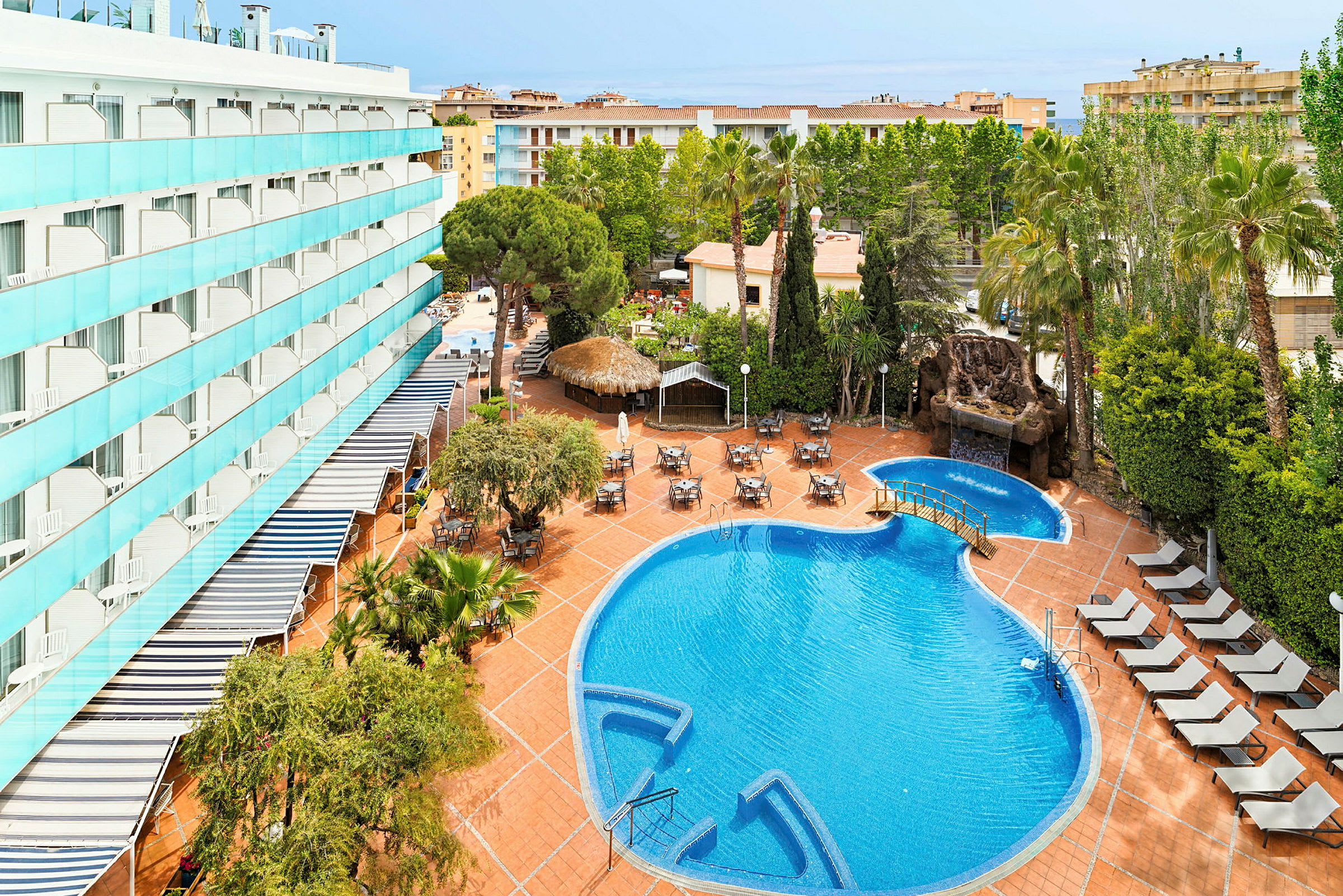 Udendørsområde med pool, liggestole og borde samt hotelbygning i baggrunden. (AI-genereret tekst)