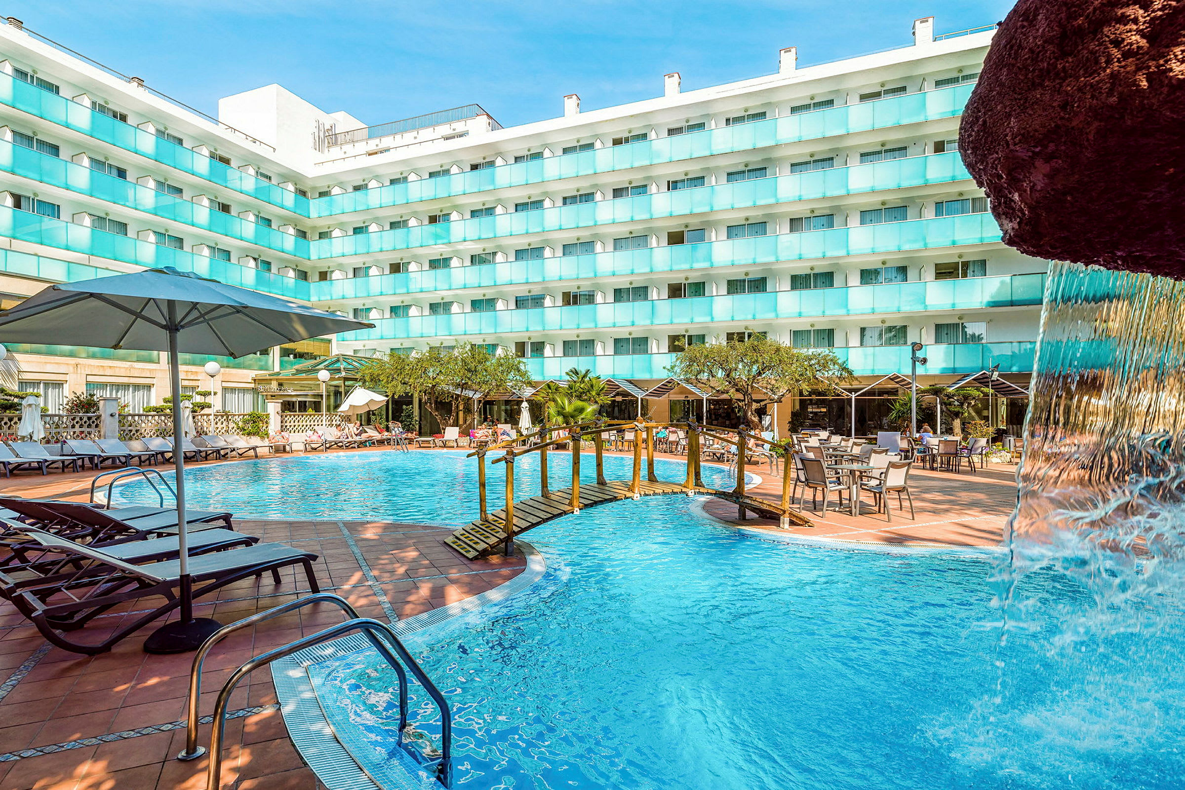 En udendørs swimmingpool ved en hotelbygning med liggestole, borde og parasoller. En lille bro over poolen og en del af en vandfaldsstruktur ses. (AI-genereret tekst)