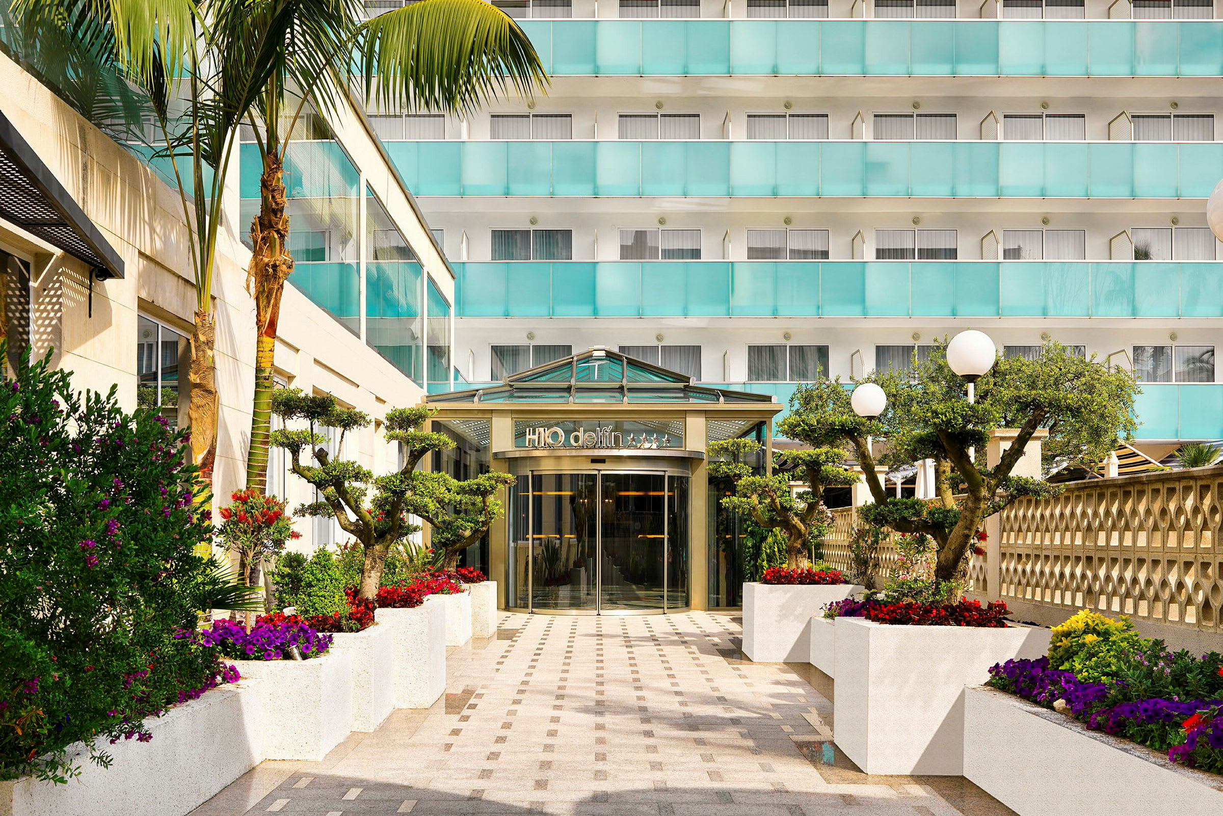 Indgang til et moderne hotel med glasdøre, omgivet af smukke blomstrende planter og træer. (AI-genereret tekst)