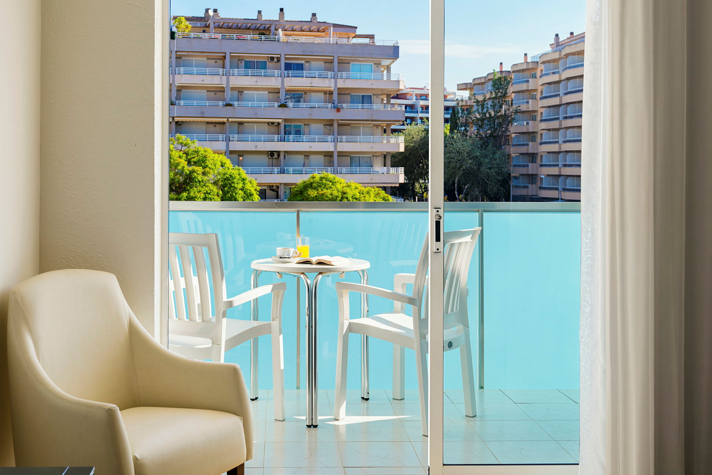 Billede af en balkon med to hvide stole og et bord med en kop og juice. I forgrunden ses en lys lænestol. (AI-genereret tekst)