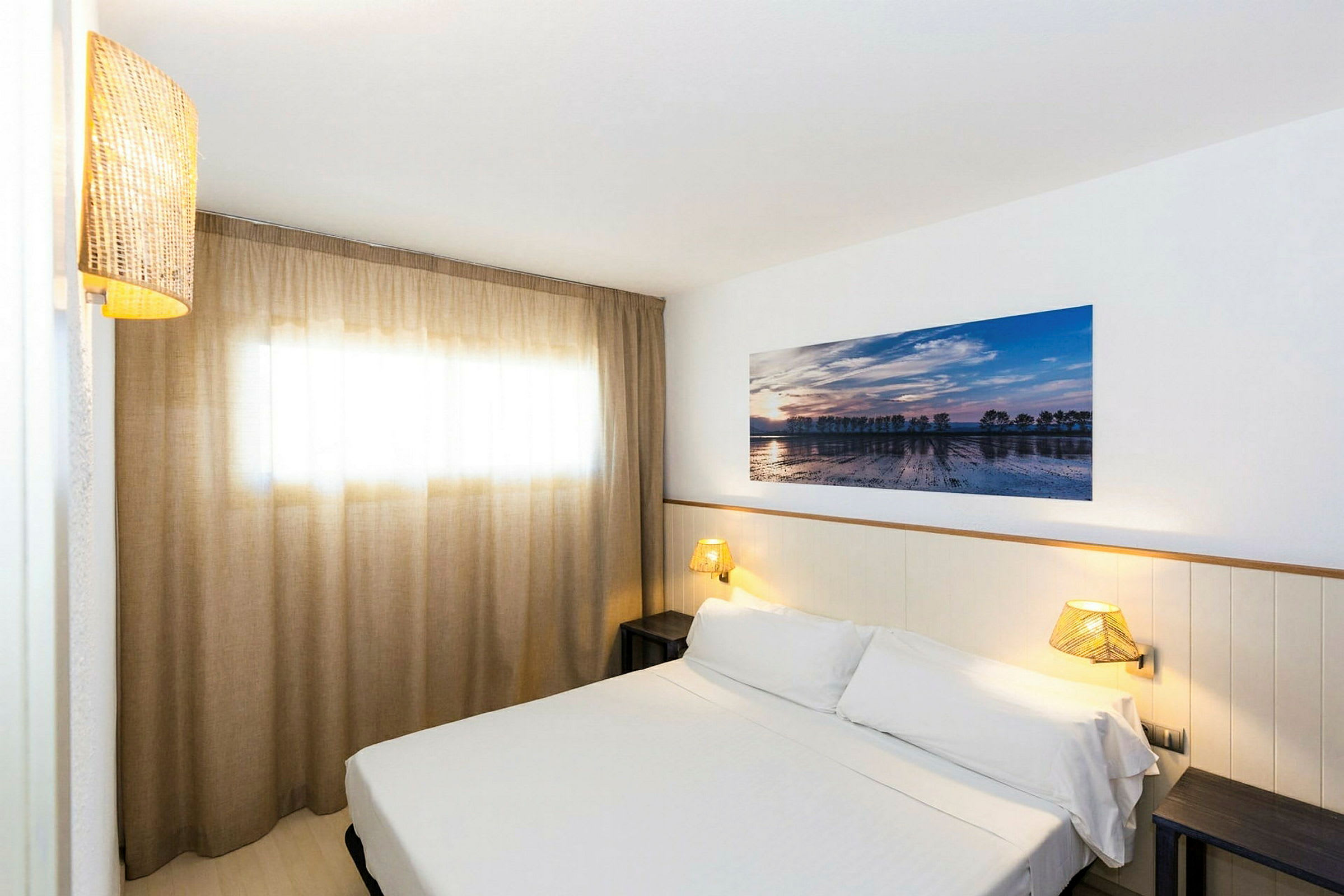 Et hotelværelse med en seng dækket med hvidt sengetøj, en væglampe og et kunstværk på væggen. Et vindue med elegante beige gardiner. (AI-genereret tekst)