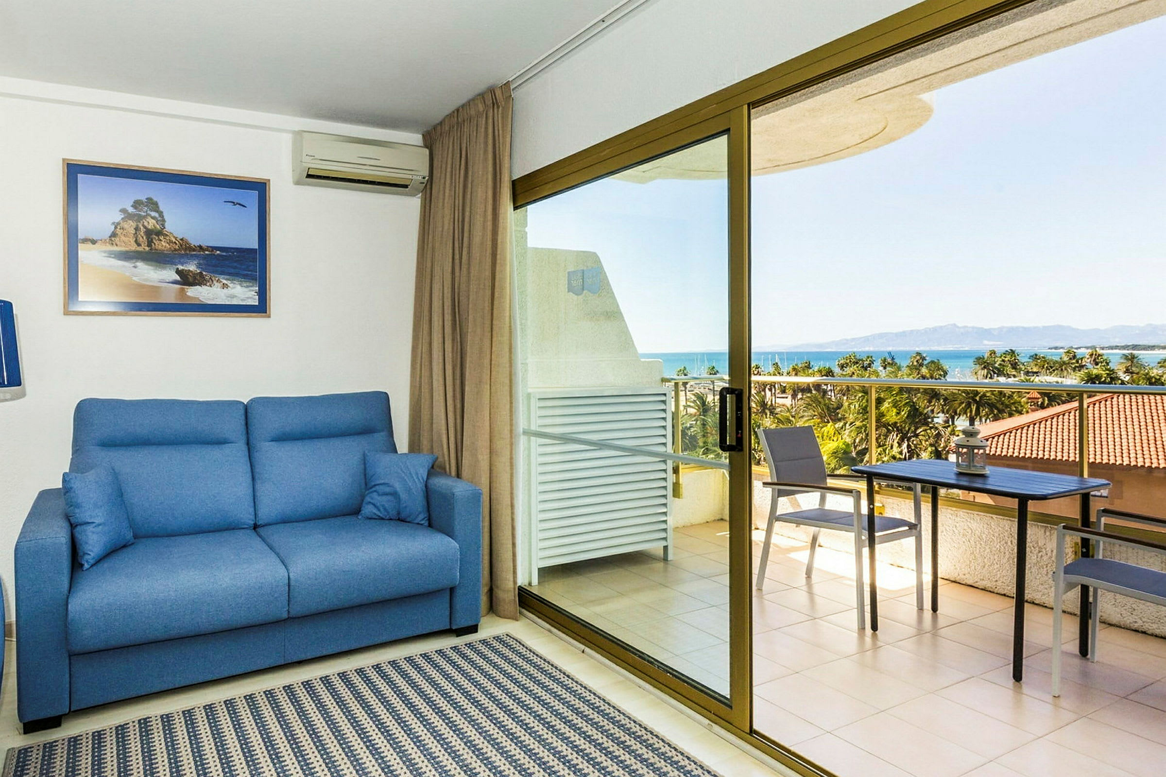 Et værelse med en blå sofa, tæppe og et glastagvindue ud mod en terrasse. Terrassen har bord, stole og udsigt over havet. (AI-genereret tekst)