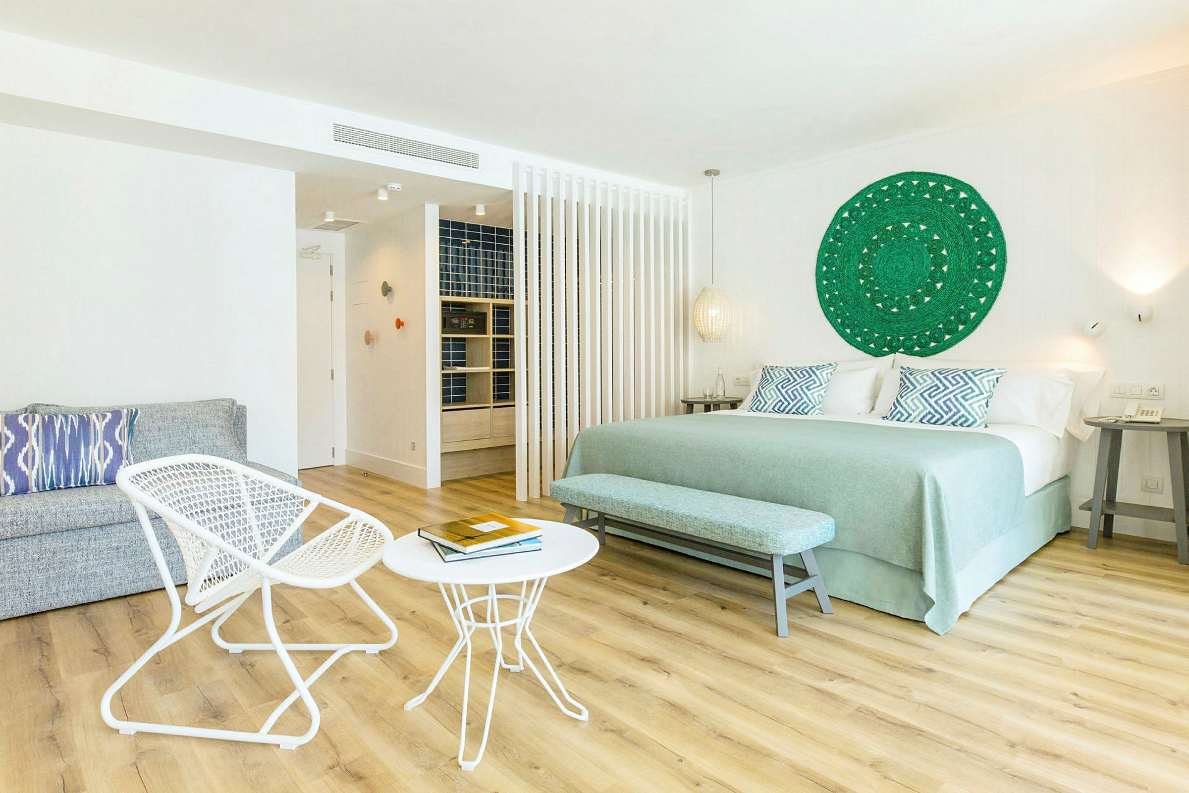 Et lyst hotelværelse med en stor seng, to små borde og to stole samt en dekorativ vægudsmykning. (AI-genereret tekst)