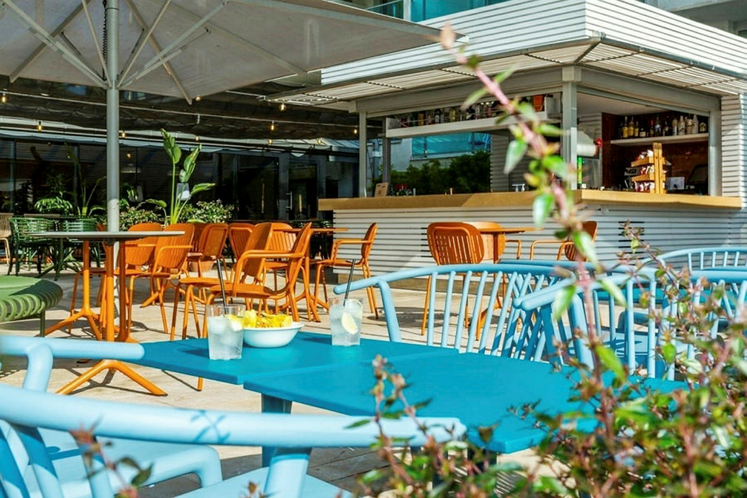 Billede af en udendørs servering med farverige stole og borde samt en lille bar i baggrunden. (AI-genereret tekst)