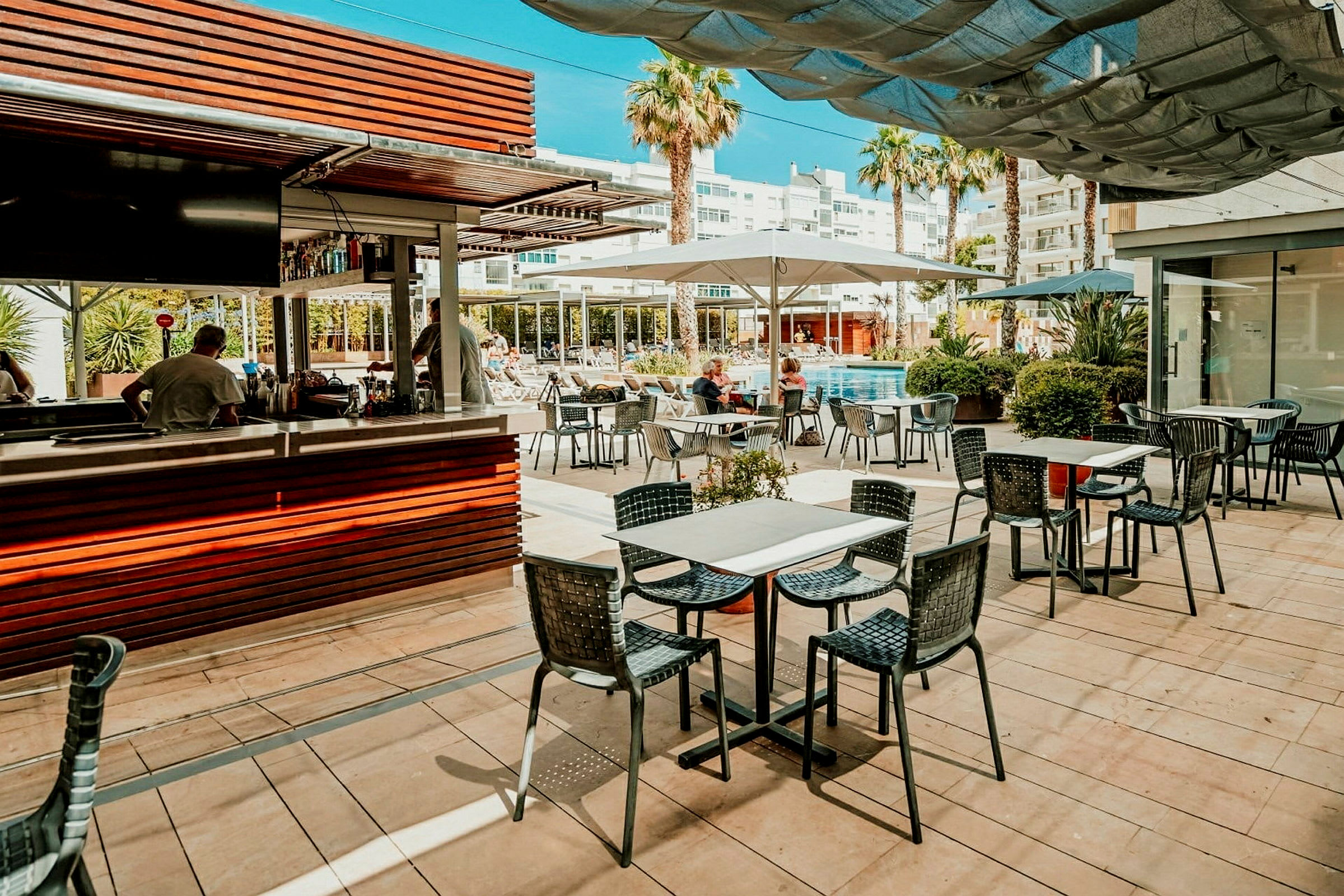 En poolnær restaurant med udendørs siddepladser og en serveringsdisk, omgivet af bygninger og palmer. (AI-genereret tekst)