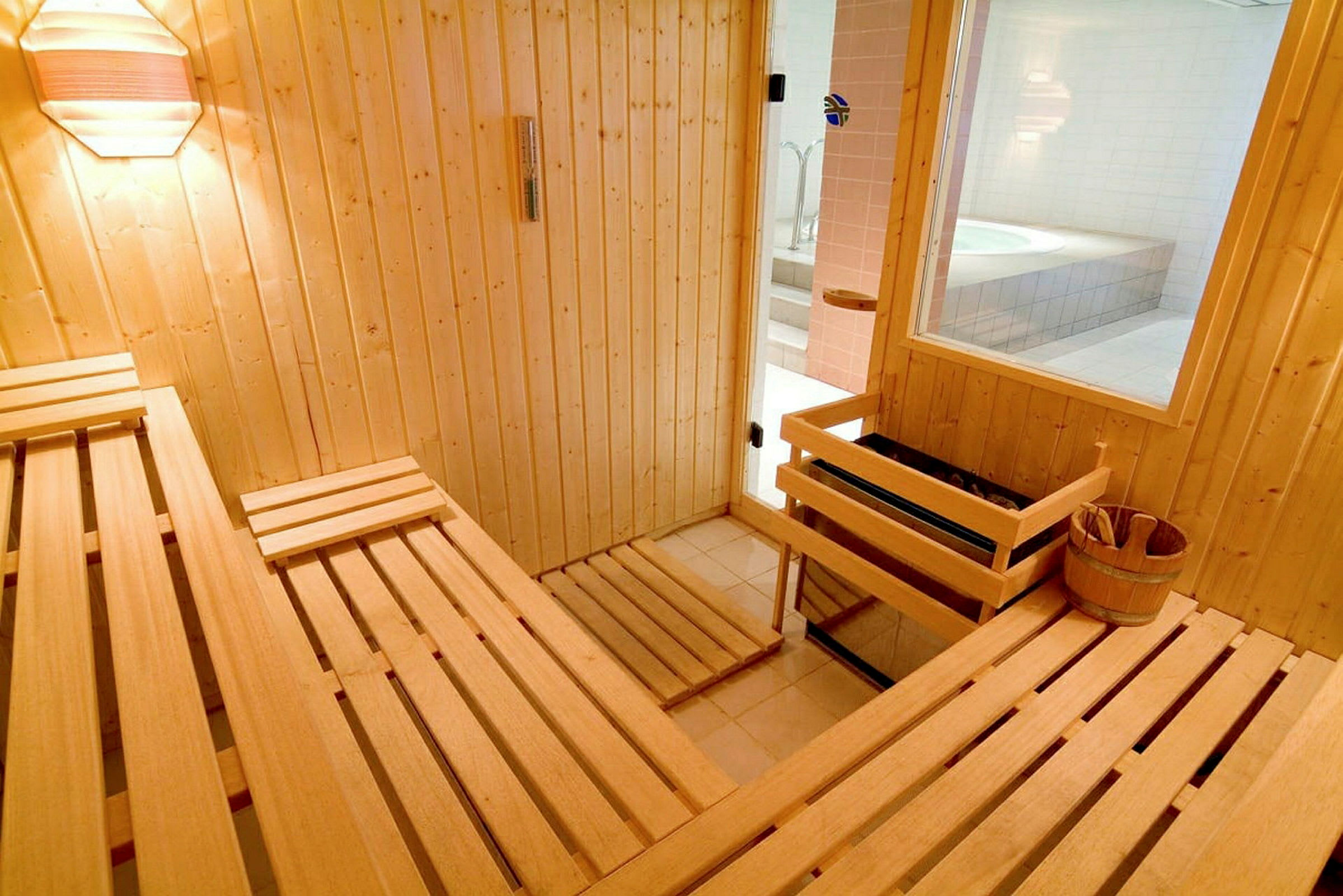 Et træinteriør i en sauna med bænke. Et vindue og en dør vender ud mod et rum med et boblebad. (AI-genereret tekst)